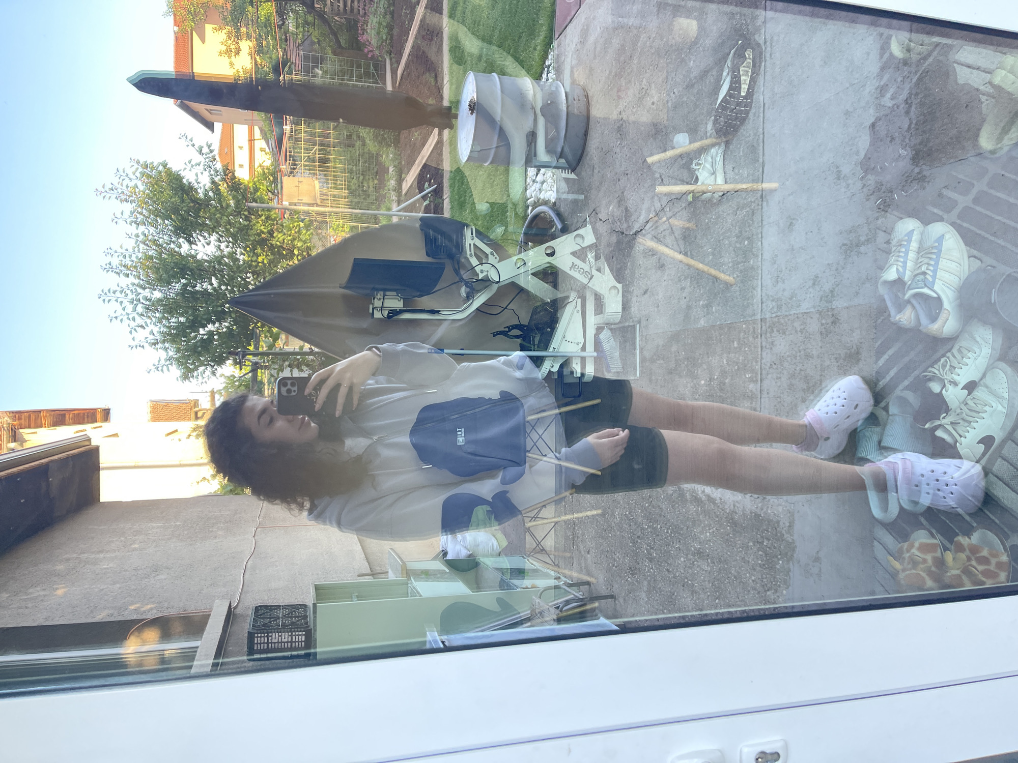 A reflection image shows a woman taking a selfie, with a background scene visible through the glass. The woman has curly hair and wears a light-colored hoodie with a dark-colored crossbody bag, black shorts, and pink Crocs. The reflected background scene includes buildings, greenery, and objects such as shoes, garden tools, a barrel, and a monitor stand. The scene may be near the city of Sofia, Bulgaria based on architectural styles and vegetation. The image is likely taken from inside a building looking outward, with the reflection creating a layered effect.
