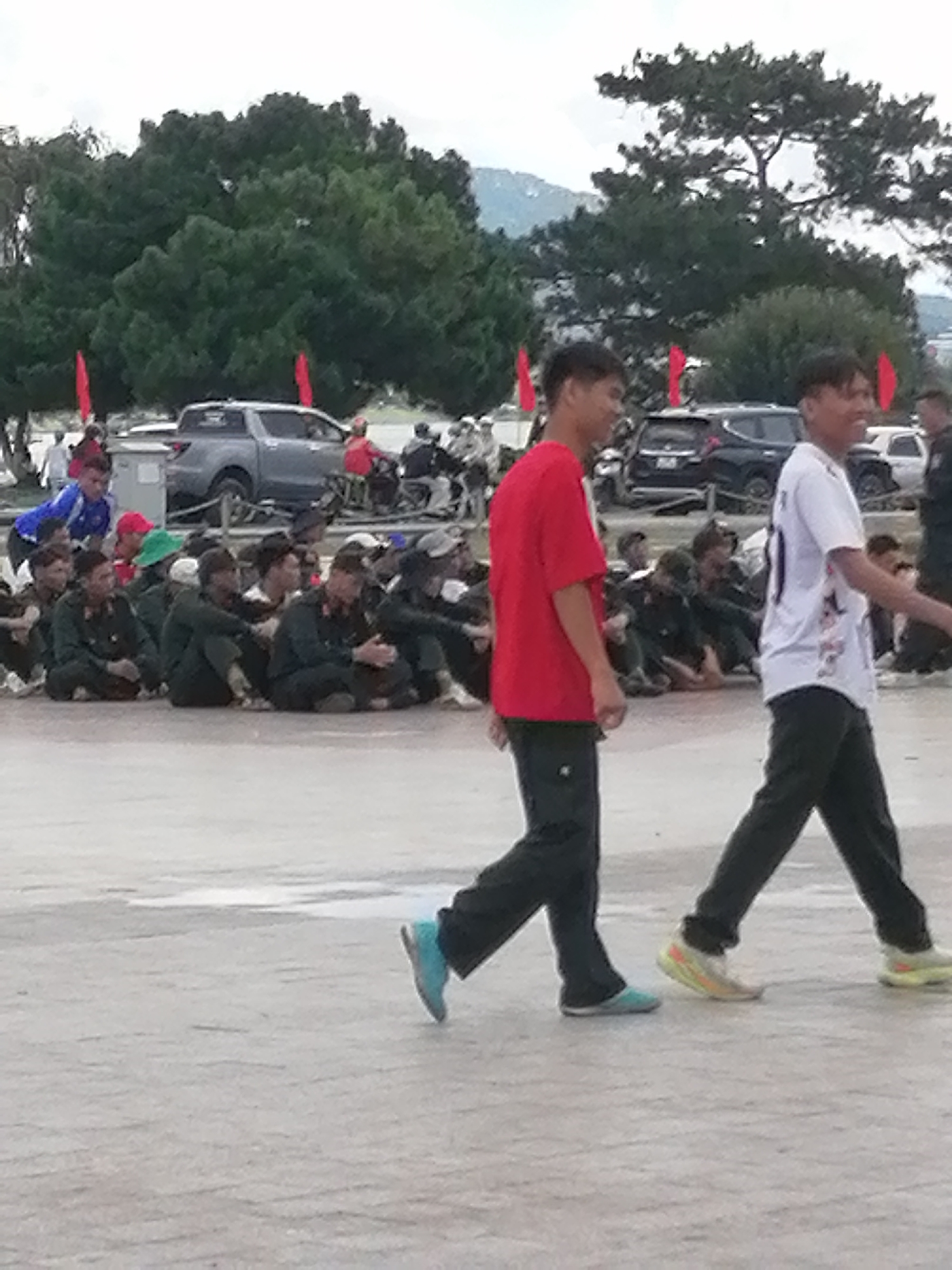 The image depicts an outdoor scene in Da Lat, Vietnam, likely near the city center, judging by the presence of a paved area, trees, and mountainous terrain in the background. In the foreground, two young men are walking towards the right. The man on the left is wearing a red t-shirt and black pants, while the man on the right is wearing a white t-shirt with black markings and black pants. Behind them, a group of individuals are seated on the ground, some appearing to be in military attire. Several vehicles, including a silver pickup truck and a dark SUV, are visible in the middle ground. Red flags are also present, scattered throughout the scene. The environment suggests a public gathering or event, possibly a parade or a festival, given the flags and the assembled crowd. The lighting indicates it is daytime, with overcast skies.