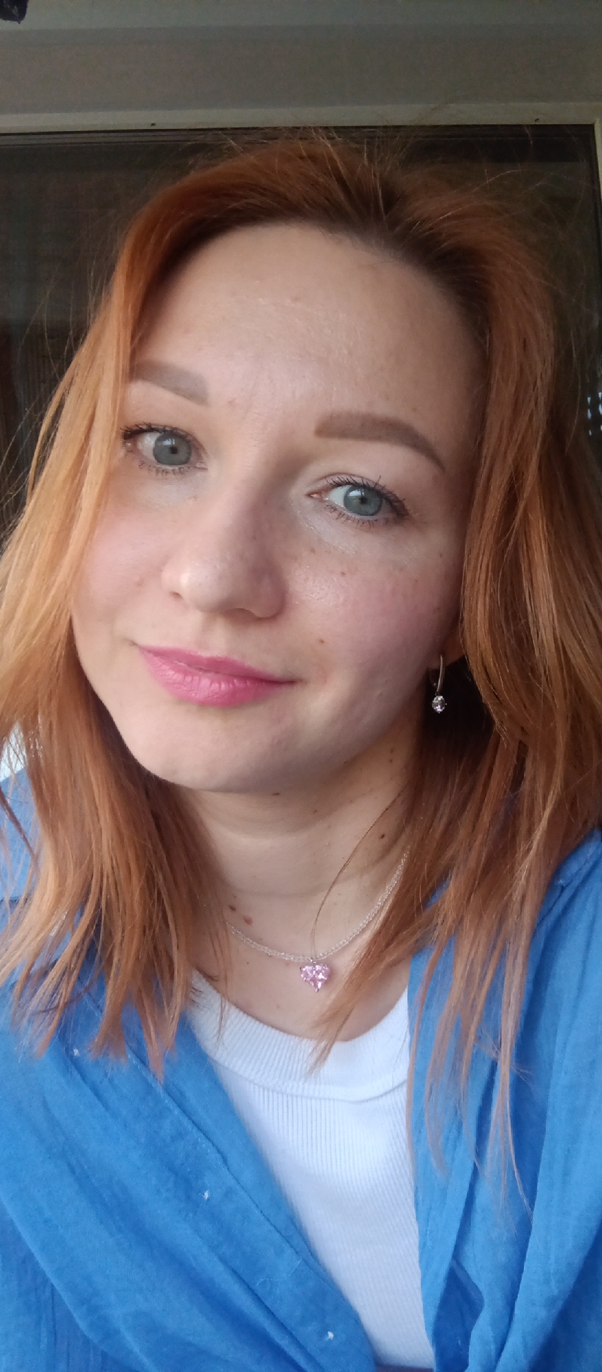 A close-up headshot of a fair-skinned woman with red hair. She is wearing a white ribbed top under a blue top, and a delicate silver necklace with a pink heart-shaped pendant. She has light blue eyes and pink lipstick. She is also wearing small, silver earrings. The background appears to be a window or doorway, but it is not clear enough to identify the location.