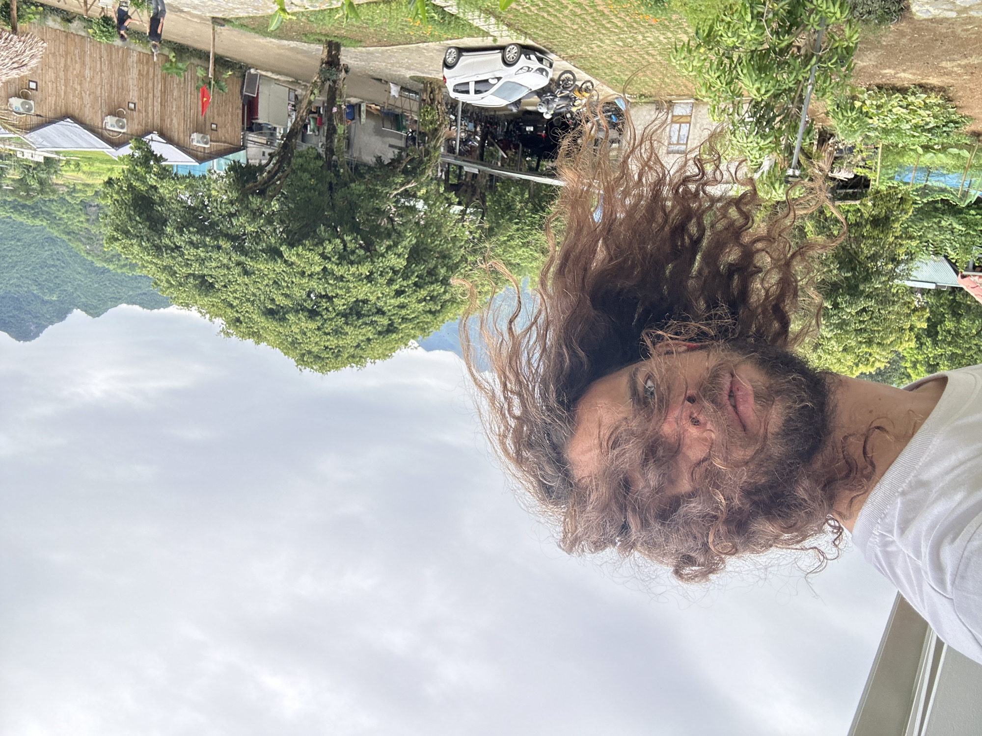 In Xóm Khán, Vietnam, a person with long, curly brown hair is leaning into the frame from the top left, upside down, wearing a white t-shirt. The person's face is visible, with blue eyes and a light beard. The background shows a rural landscape with lush green trees, mountains in the distance, and a cloudy sky. In the middle ground, there are several bamboo-style buildings with thatched roofs, air conditioning units visible on the walls, and a Vietnamese flag flying. A white car is parked on a paved area in front of one of the buildings, alongside several motorcycles. Further left, there are more buildings, some with corrugated roofs, and a vegetable garden. Two individuals, a man and a woman, are walking on a pathway in front of the bamboo buildings. The scene appears to be an outdoor area, possibly a guesthouse or a small village.