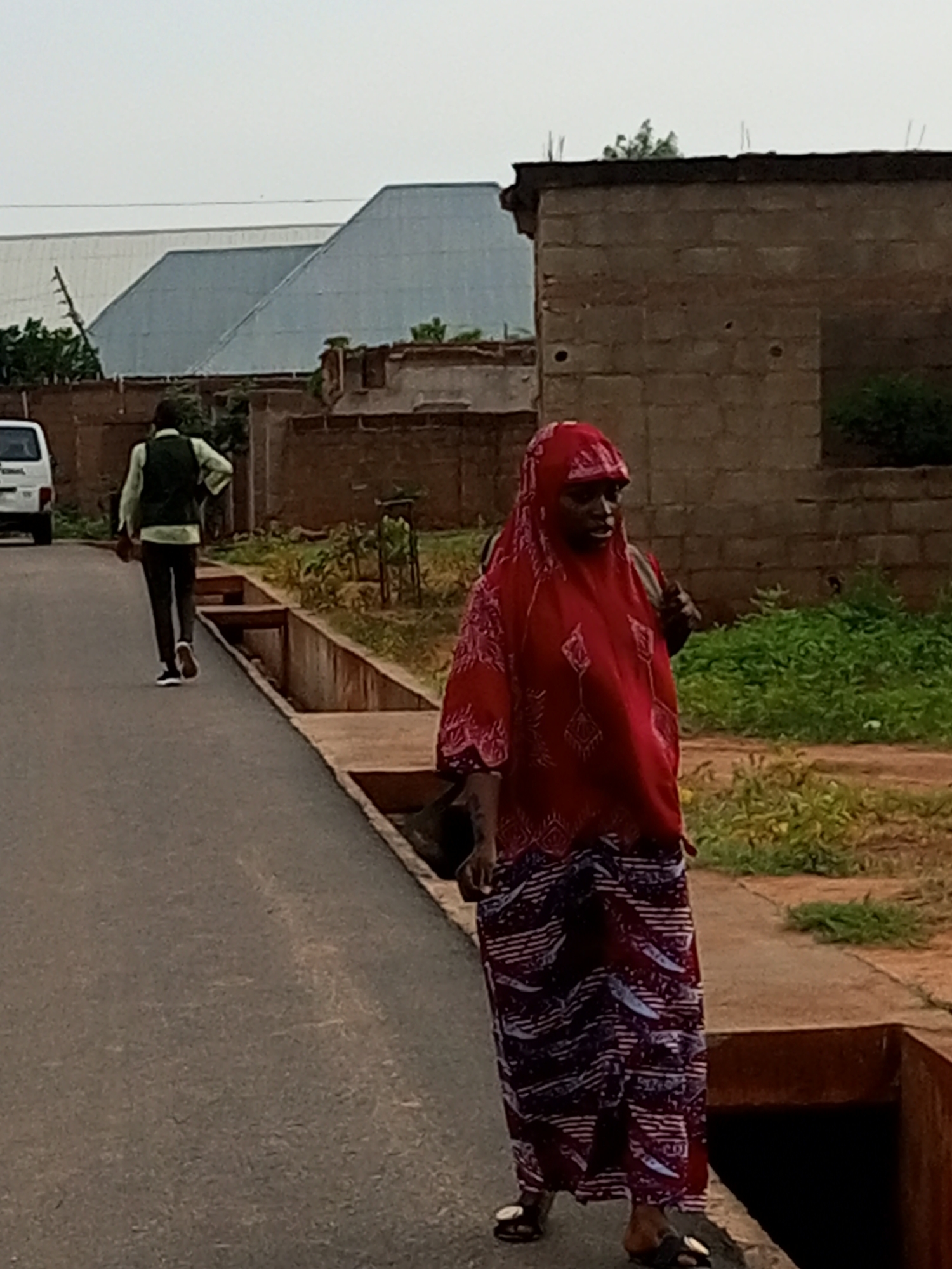 In Tempure, Nigeria, an outdoor scene unfolds on an asphalt road. A woman wearing a red hijab and a patterned red and white dress walks towards the viewer, her feet shod in black sandals with gold accents. She carries a dark bag over her left shoulder. Behind her, a man in a light-colored shirt and dark pants, also wearing a vest, walks away from the camera. He is dressed in dark sneakers. The road is bordered by a concrete channel on the right, with overgrown vegetation beyond it. In the background, a brick building with a flat roof stands to the right, while a structure with a corrugated metal roof is visible to the left, partially obscured by trees. A white van is parked on the left side of the road. The sky is overcast.