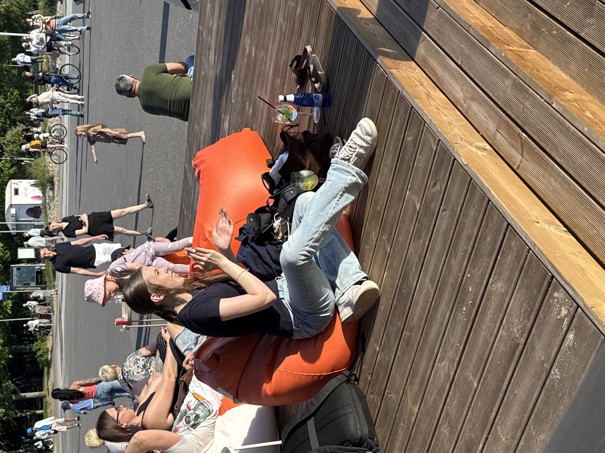 In Tallinn, Estonia, an outdoor scene unfolds on a wooden deck under a bright, sunny sky. A young woman, seated on a large orange beanbag chair, is the focal point, her arms raised and hands clasped as if clapping. She wears a dark t-shirt and light blue jeans. Nearby, a backpack rests on the deck. On the wooden surface, two glasses with drinks, one containing ice and mint, and a blue bottle of liquid are visible. A pair of flip-flops lie to the right. In the background, other individuals are scattered, some walking, others seated, and some on bicycles, suggesting a public park or recreational area. The setting appears to be a waterfront or a promenade.
