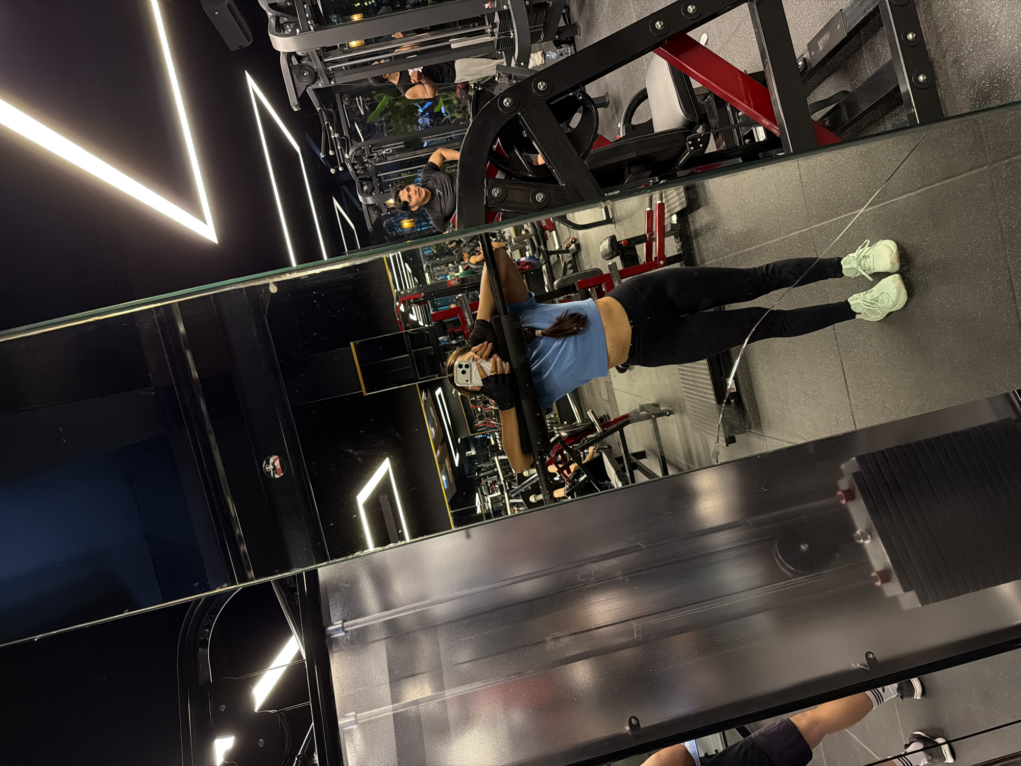 The image shows a woman in a gym, reflected in a large mirror. She is wearing a light blue shirt, black leggings, and light green sneakers. She is holding a black bar above her head and looking at her phone, likely taking a photo. The gym has modern lighting, black walls, and various exercise machines, including a leg press. Another person, a man wearing a dark t-shirt, is visible in the mirror, standing near the gym equipment. The location is likely in Denpasar, Indonesia, though this is not directly evident in the image.