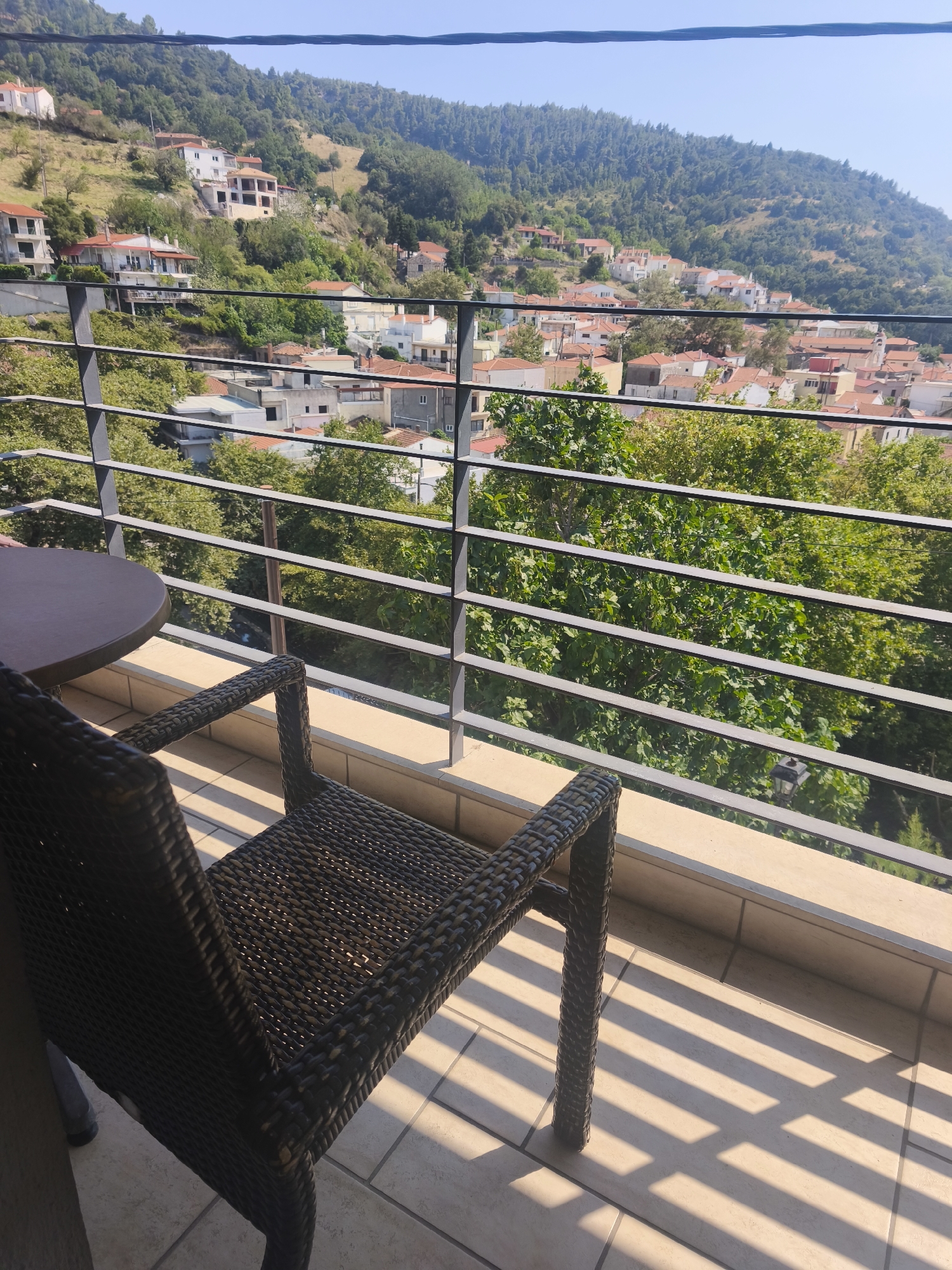 The image captures a view from a balcony in Στενή Δίρφυος, Greece. In the foreground, a dark woven chair is positioned on a tiled balcony, with its legs casting long, striped shadows on the floor due to sunlight filtering through a railing. A dark round table is partially visible to the left of the chair. The balcony railing, made of dark metal bars, separates the viewer from the landscape beyond.
Beyond the railing, a dense canopy of green trees dominates the middle ground. In the background, a hillside settlement is visible, characterized by numerous buildings with red-tiled roofs, interspersed with more greenery. The settlement appears to be built on a slope, with houses cascading down the hillside. The sky above is a clear, bright blue. The overall scene depicts a tranquil, scenic view of a Greek village nestled in a verdant, hilly landscape.