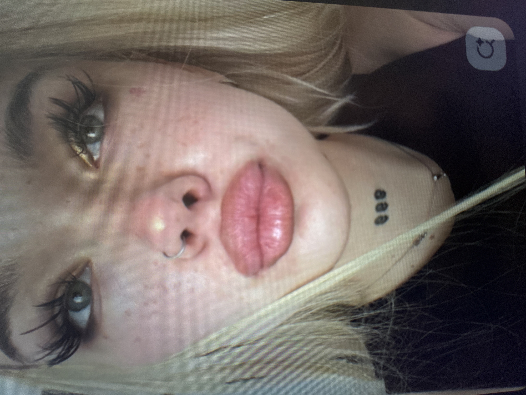 A close-up, rotated image captures the face of a person, likely a young woman, with light blonde hair framing her face. Her head is tilted, with her eyes looking towards the camera. She has light-colored eyes, possibly green or hazel, accentuated by long, dramatic false eyelashes and dark eyeliner. Her complexion is fair with visible freckles across her nose and cheeks.
Her nose features a silver hoop nose ring in the right nostril. Her lips are full and pink, appearing prominently in the center of the frame, possibly enhanced. On the right side of her neck or jawline, a small tattoo of three vertically aligned black dots is visible, along with what appears to be a piercing or piece of jewelry.
The background is dark and indistinct, suggesting an indoor setting or low-light conditions. The image itself appears to be a photograph of a screen, indicated by visible pixelation and a user interface icon in the top right corner. This icon depicts a refresh or rotation symbol, incorporating a stylized smiley face with stars for eyes. The person maintains a neutral to slightly pouty expression.
