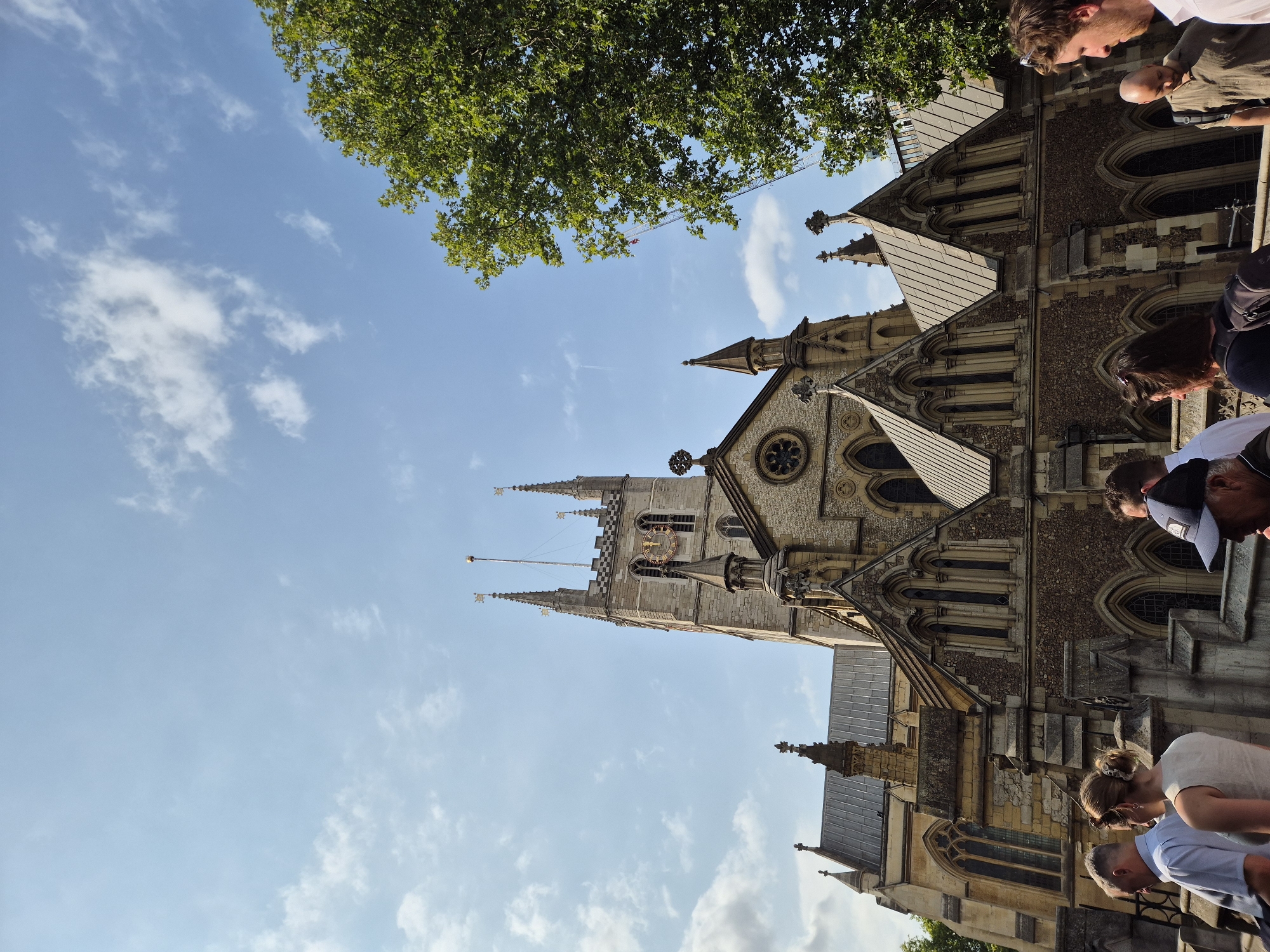 The image shows a low-angle shot of Southwark Cathedral in London, United Kingdom, under a bright blue sky with scattered white clouds. The facade of the cathedral is made of stone and features Gothic architectural elements, including pointed arches, spires, and a large rose window. A clock face is visible on the central tower. Several people are walking in the foreground, some looking towards the cathedral. A large green tree with dense foliage is visible on the right side of the image, partially obscuring the cathedral. The overall scene suggests a sunny day with tourists or visitors exploring the historic landmark.