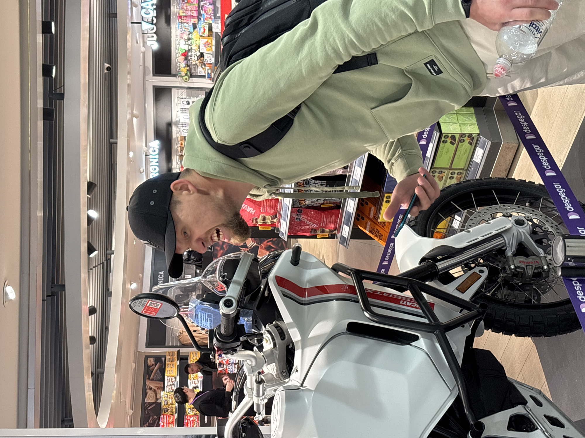 This is a candid, vertical photograph taken indoors, likely in a retail environment such as an airport or a large store in Pudahuel, Chile. The main subject is a young man, possibly in his late twenties or early thirties, wearing a light green hoodie and a black baseball cap. He is leaning over and looking at a white motorcycle. His mouth is open, as if he is speaking or expressing surprise, and he is holding a green pencil in his right hand. He is also holding a partially empty plastic water bottle in his left hand.
The motorcycle appears to be a Ducati, with prominent "DUCATI" and "DESERTX" branding visible. It is white with red and grey accents on the fairing. The front wheel and part of the suspension are visible.
In the background, there are shelves stocked with various products, including what appear to be chocolates and other food items. Some of the packaging displays text such as "HAVANNA" and "CACAO PURO". There are also purple banners with white text that reads "despegar". The lighting suggests it is daytime, and the environment appears to be well-lit and modern. The overall mood is casual and engaging, with the man seemingly interacting with the motorcycle or someone out of frame.
