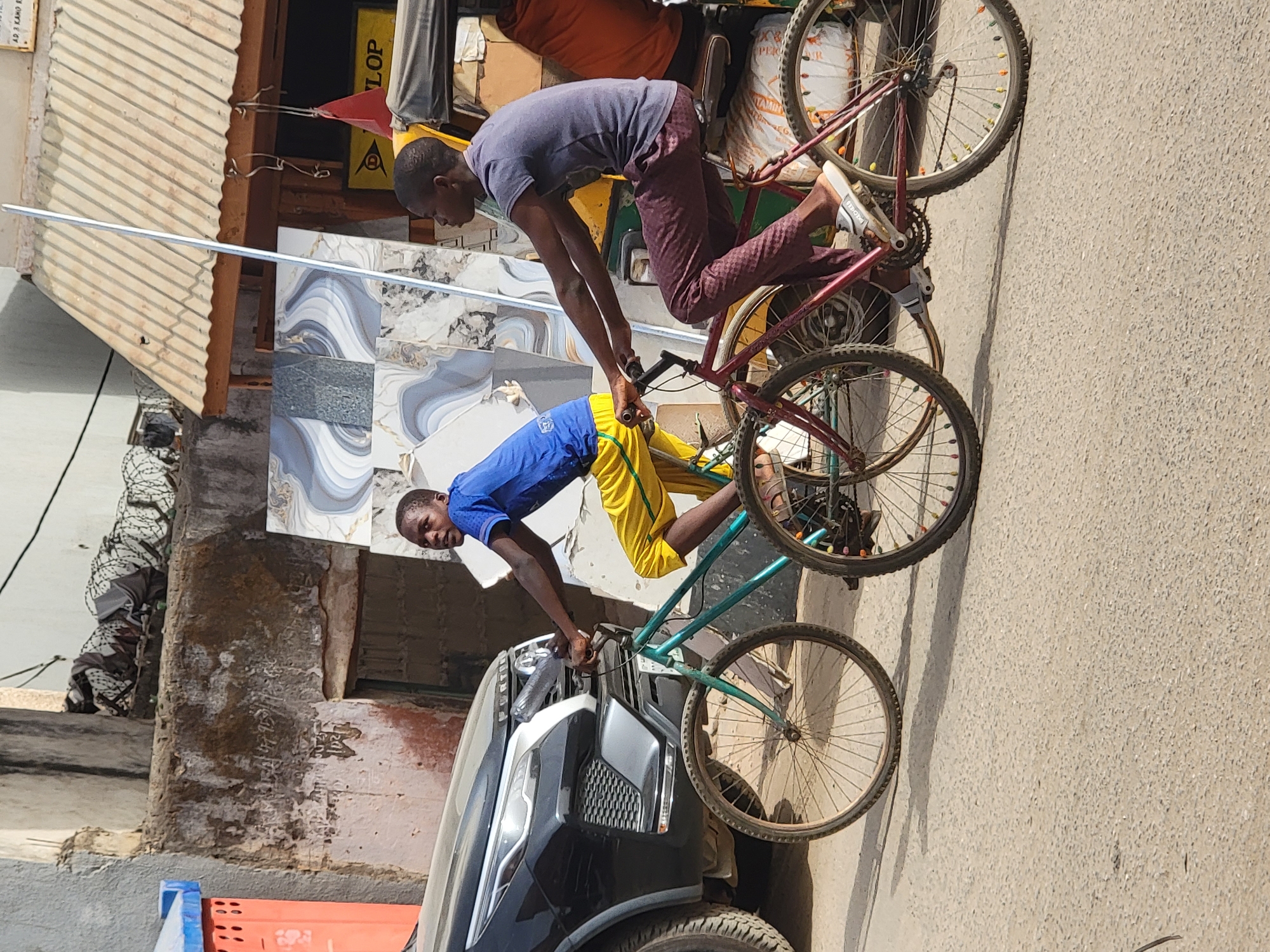 Two young men are engaged in a balancing act with bicycles on a sunny day in what appears to be an urban or semi-urban street in Maiburiji, Nigeria. The scene is illuminated by bright daylight, casting strong shadows on the ground.
In the foreground, a young man wearing a grey-purple t-shirt and patterned dark pants is balanced precariously on the frame of a red bicycle. He is crouched forward, gripping the handlebars with both hands, his focus seemingly on maintaining his stability. His light-colored shoes are visible on the pedals or frame. The red bicycle has colorful beads decorating its spokes.
Beside him, another young man in a blue t-shirt and yellow shorts is holding the handlebars of a green bicycle. He is looking towards the viewer with a slight smile, suggesting he is part of the performance or aware of being photographed. This green bicycle also has spoked wheels. The front of a black SUV or car is partially visible behind the man in blue.
In the background, there is a building with a corrugated metal roof and a wall featuring decorative blue and white patterned tiles. A small part of another person, dressed in patterned clothing, is visible on the far left. Various objects like bags and signs are scattered in the background. Partial text is visible on a yellow sign, showing "LOP", likely from "DUNLOP", and on a white bag, "VITAMIN". Another sign shows "AD I KAN" and "R". The ground is a light-colored, somewhat dusty surface, possibly a road or pathway.