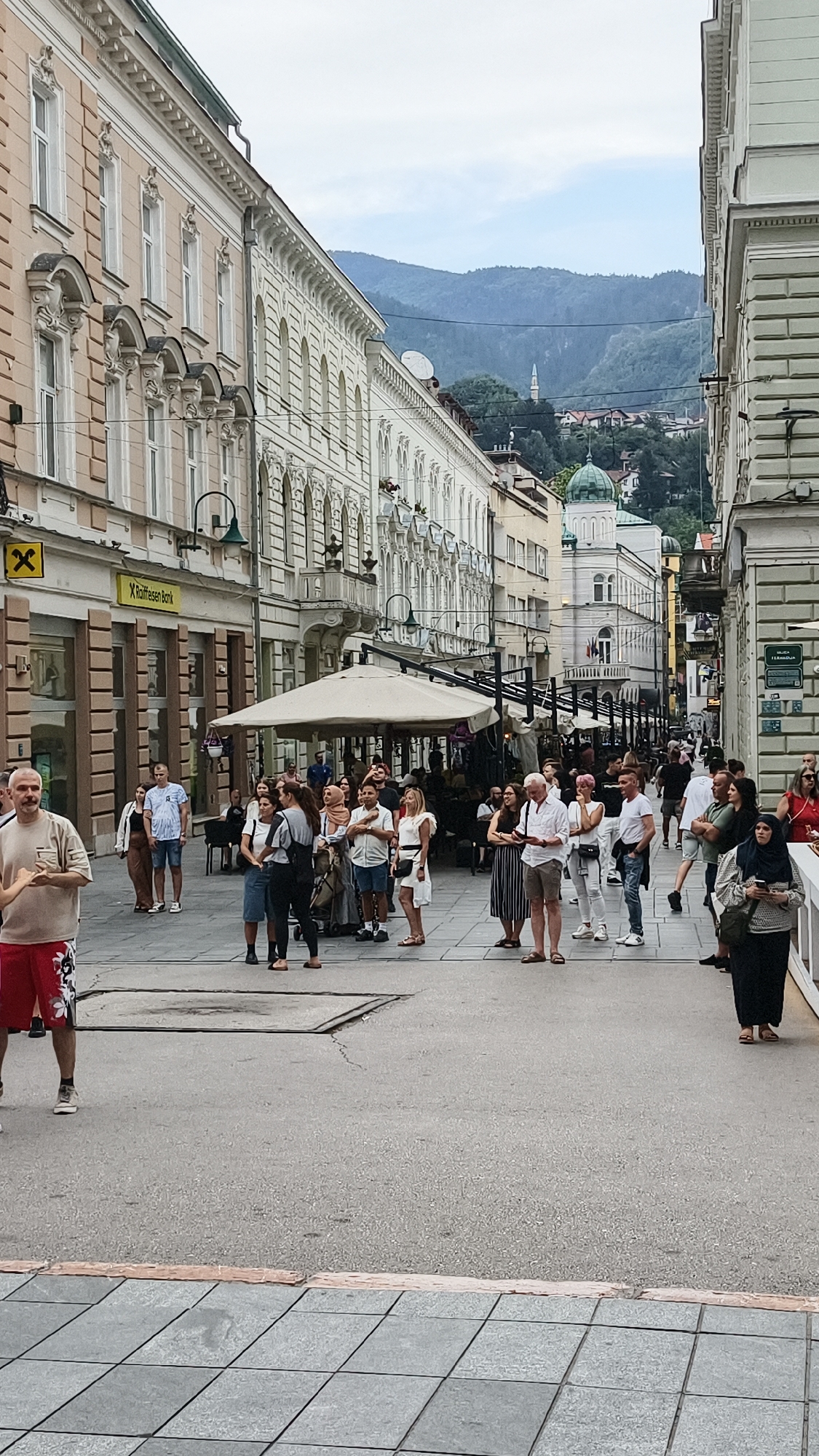 The image captures a bustling pedestrian street in Sarajevo, Bosnia and Herzegovina, likely in the city center. The architecture features ornate, light-colored buildings with decorative facades, indicative of Austro-Hungarian influence. A Raiffeisen Bank sign is visible on one of the buildings. The street is lined with outdoor seating for cafes or restaurants, shaded by large white umbrellas. A diverse crowd of people, dressed in casual summer attire, are milling about, some standing and talking, others walking. The ground is paved with asphalt in the main thoroughfare and grey tiled sections. In the background, a mosque minaret and other buildings are nestled against a backdrop of lush, green hills and mountains under a cloudy sky. The overall scene depicts a vibrant urban environment with a mix of historic architecture and contemporary life.