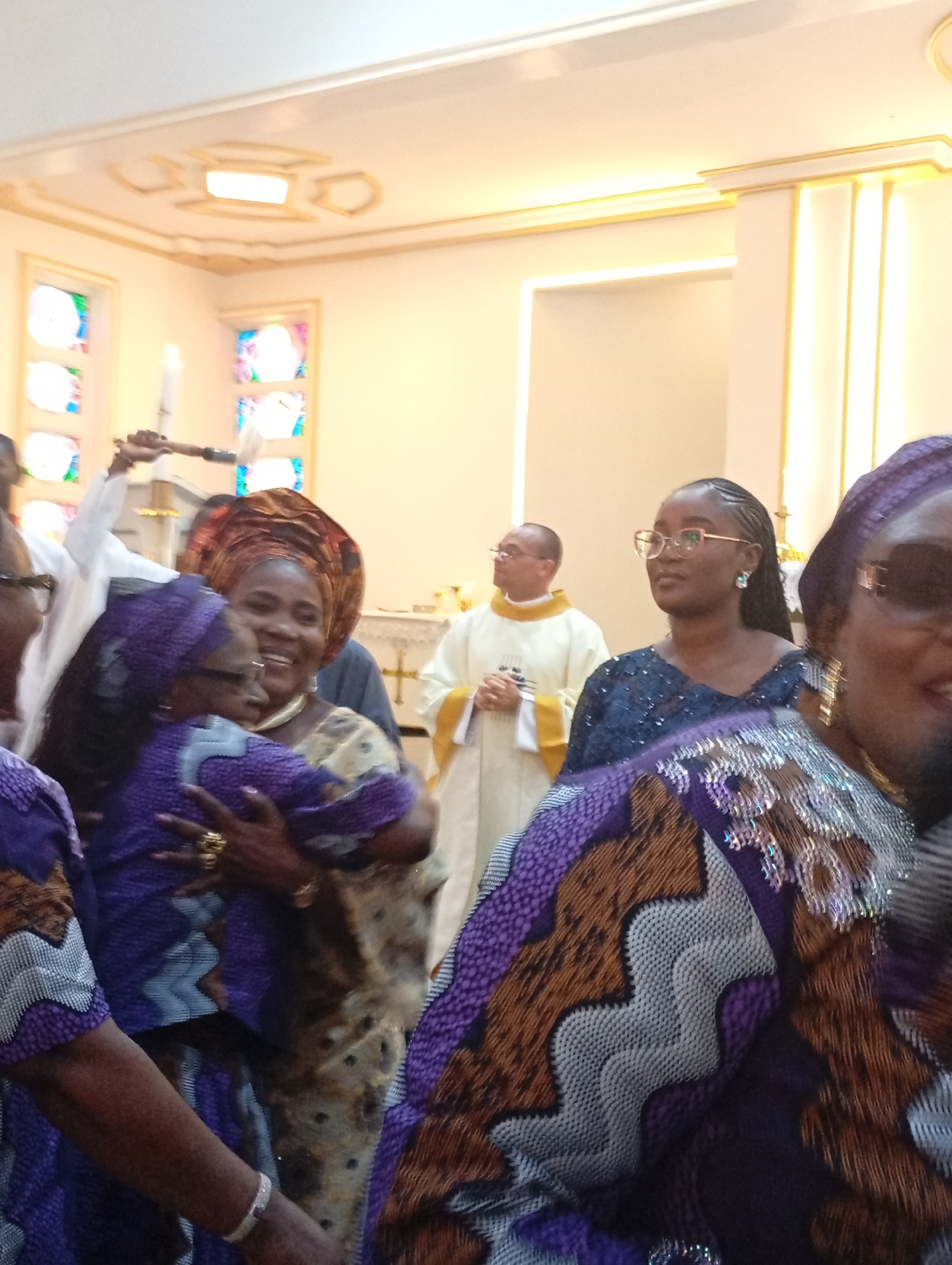 The image depicts an indoor scene, likely a church or chapel, located in an unknown city. Several individuals are present, including women in patterned traditional clothing, a priest wearing a white robe, and another person holding a candle with a brush or similar implement. An older woman is seen embracing another woman, both wearing similar patterned garments. The church's interior features light-colored walls with gold trim, stained glass windows, and ornate ceiling light fixtures.