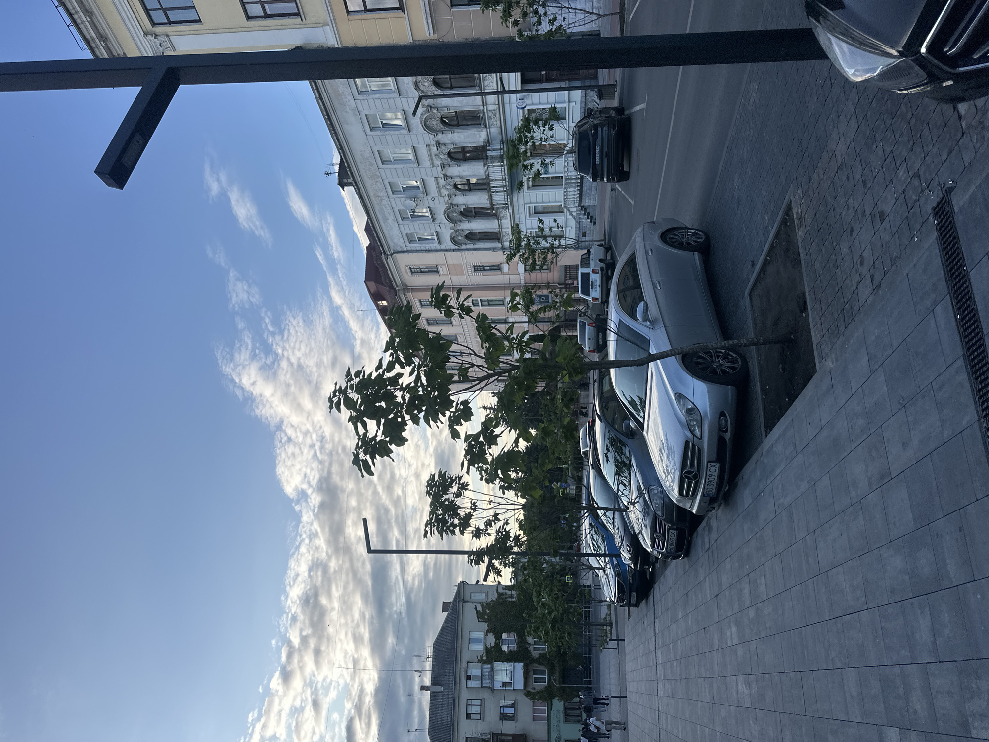 The image depicts a street scene in Ternopil, Ukraine, at what appears to be either sunrise or sunset, given the lighting conditions and the presence of clouds in the sky. The foreground is dominated by a paved sidewalk and a street with several cars parked along the side.
On the sidewalk, a few trees are planted in square plots. The paving stones are a dark grey color, laid out in a regular pattern. A modern, black street lamp stands tall on the right side of the frame, its design minimalist and angular.
The street itself is also paved, and several vehicles are visible. From left to right, the cars include a blue sedan, a dark gray hatchback, a silver sedan (likely a Mercedes-Benz, with license plate ending in CX), and a black SUV. Further down the street, a white SUV and another dark-colored SUV are parked.
In the background, there are several buildings. The closest building on the right is light yellow with cream-colored trim, featuring classical architectural elements like columns and decorative moldings above the windows. To the left of this building is a lighter-colored building with arched windows on the second floor and a balcony. Further to the left, a multi-story residential building with a grey facade and balconies is visible.
A few individuals are walking on the sidewalk in the distance, near the residential building. Their presence is subtle and does not appear to be the primary focus of the scene. The sky is a mix of blue and white, with some scattered clouds, suggesting mild weather. The overall impression is that of a quiet urban street with parked cars and some architectural interest from the historical buildings.