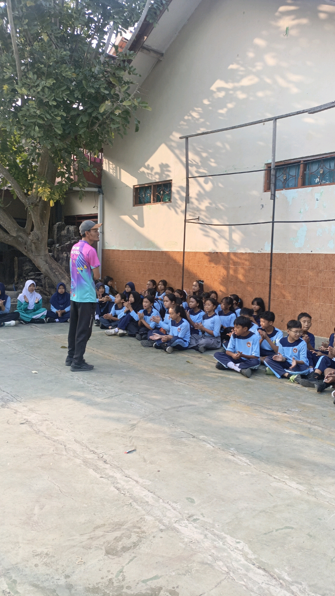 A group of young students, predominantly female, wearing blue and white uniforms are sitting cross-legged on a concrete ground in an outdoor setting. They appear to be attentively listening to a male instructor, who is standing and facing them. The instructor is wearing a colorful t-shirt with a design on the front, dark pants, and a baseball cap. Some of the students are clapping their hands.
In the background, there is a building with a tiled roof and light-colored walls. Two windows with decorative grills are visible on the wall. A large tree with dense foliage is situated to the left of the scene, casting shadows on the ground and the building. The lighting suggests it is daytime, possibly morning or late afternoon. The ground surface is cracked concrete.
This scene is located in the city of Salatiga, Indonesia.