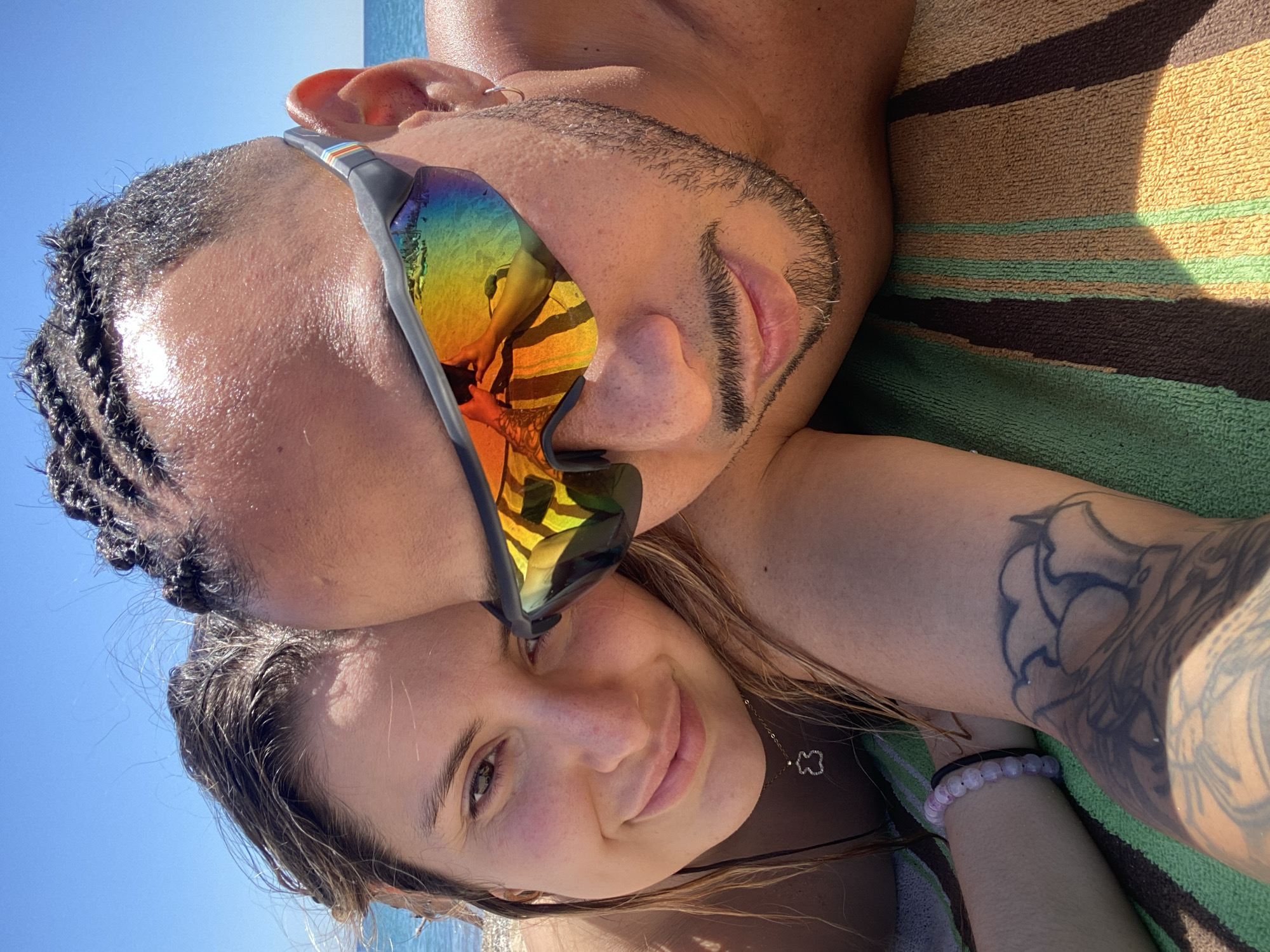 A close-up shot depicts a light-skinned man and a light-skinned woman lying together on a striped beach towel against a backdrop of clear blue sky and water. The man is positioned above the woman, his face close to hers. He is wearing sunglasses that reflect a beach scene with colorful stripes. His hair is styled in braids, and he has a short beard. The woman is smiling. She is wearing a necklace with a small pendant, and a light purple bracelet. She has a tattoo on her right arm. The setting suggests a beach location, potentially in Barcelona, Spain, though specific landmarks are not visible to confirm the precise location.