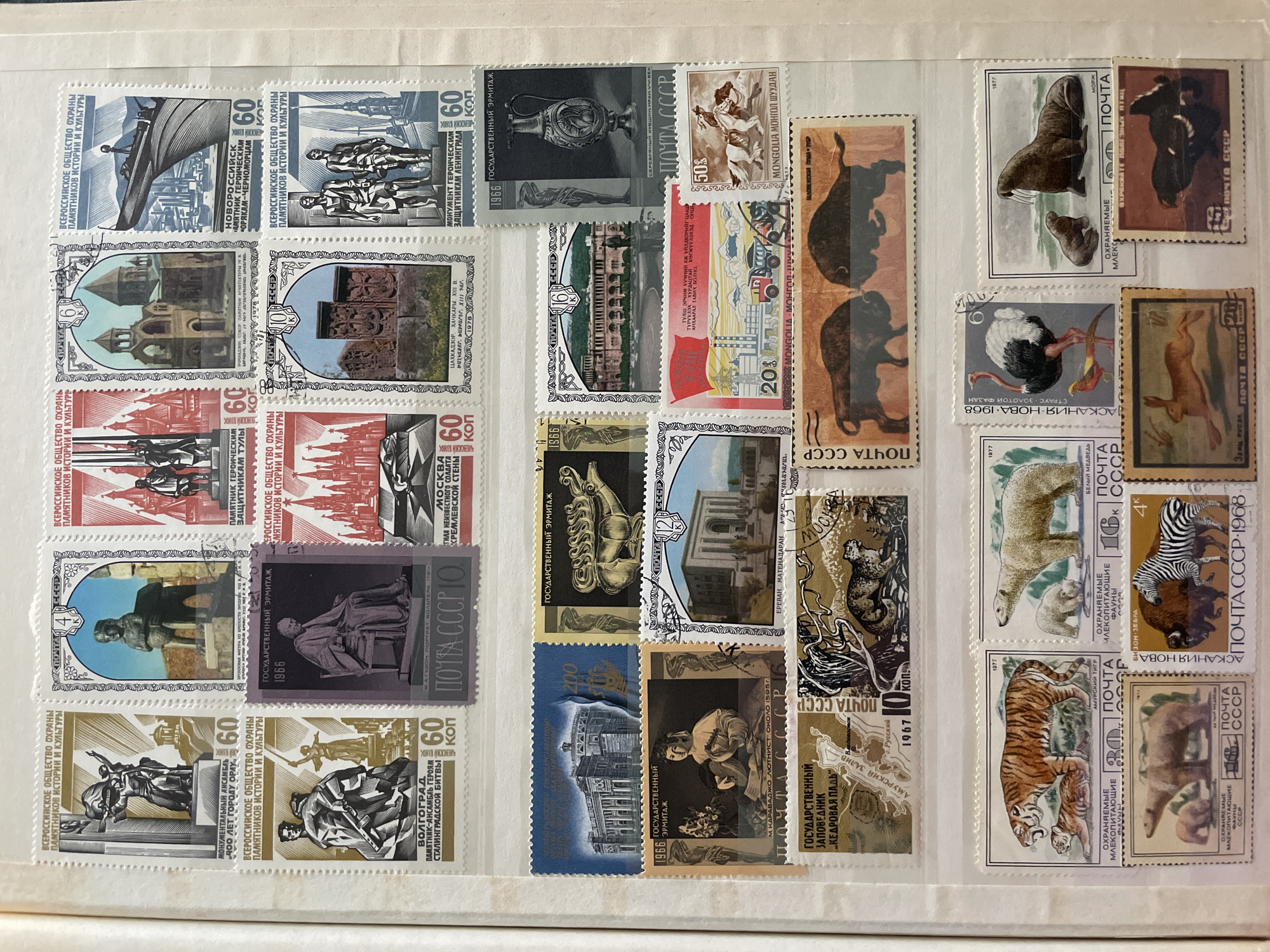 The image displays a collection of postage stamps. The stamps feature various historical monuments, architectural buildings, and wildlife. Several stamps are from the USSR, with inscriptions such as "ПОЧТА СССР" (POST OF USSR). Other stamps appear to be from Mongolia, as indicated by "MONGOLIA" and "МОНГОЛ ШУДАН". One stamp has a red border and shows a factory with a truck and a bulldozer, with text in Cyrillic. Another stamp depicts a tiger and its cub, while a separate stamp shows a polar bear with its cub. A stamp features an ostrich and a pheasant. Another stamp shows a walrus and its pup. Additionally, there are stamps illustrating bison and zebras, a rabbit, and a bird of prey. The stamps are arranged in rows and columns on a light-colored background, suggesting they are part of a collection album. No specific location in the city of Сокол, Russia, is depicted or referenced in the image.