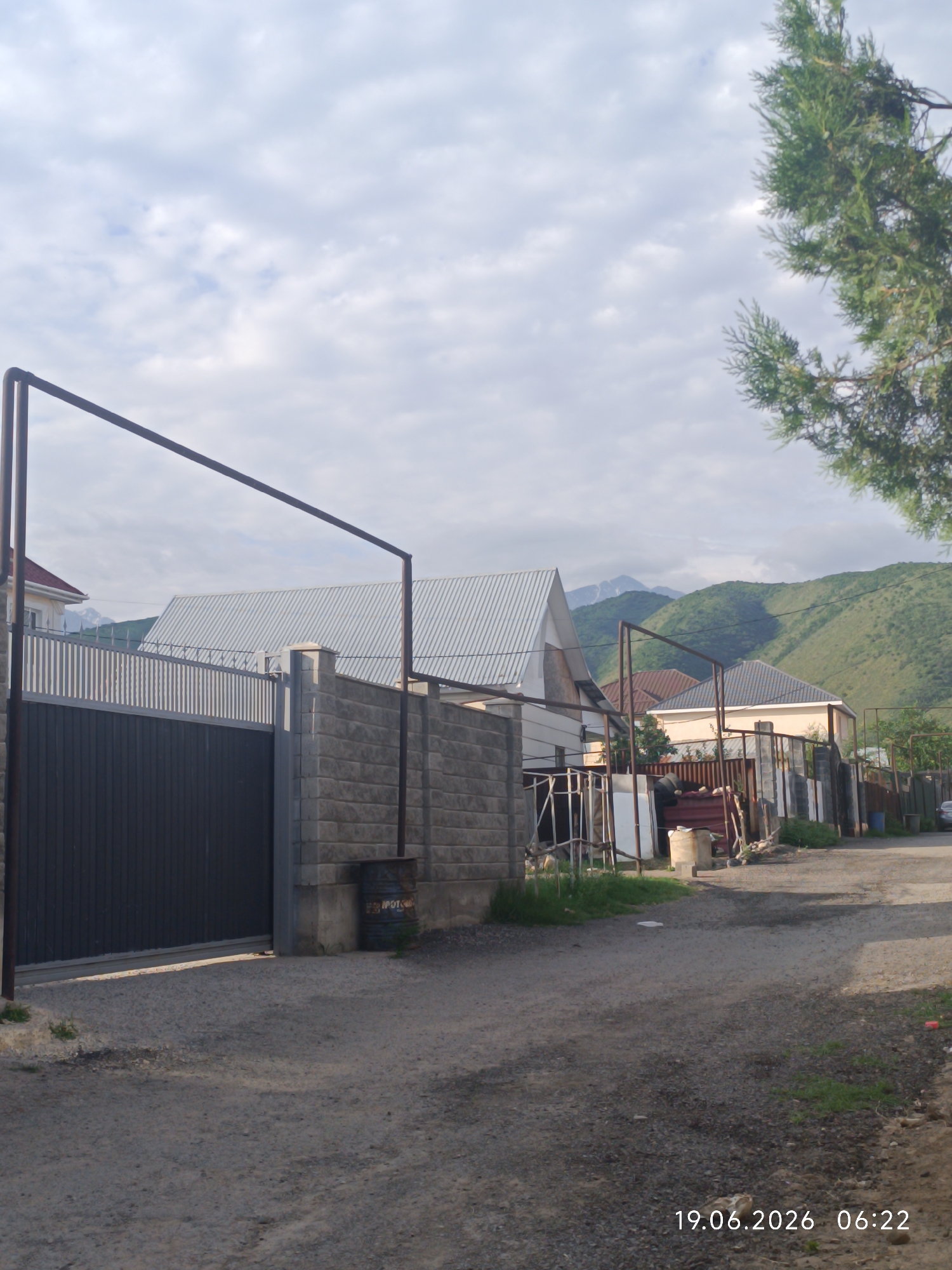 The image depicts a street scene in the city of Қырғауылды, Kazakhstan. A large, dark gray metal gate dominates the left side of the frame, flanked by a textured gray stone wall. Beyond the wall, several houses with varied roofing materials are visible, including corrugated metal and what appears to be asphalt shingles. In the background, lush green mountains rise under a cloudy sky. A large green tree with dense foliage partially obscures the upper right portion of the image. The ground in the foreground is a gravel road, with some sparse grass growing along the edges. A rusted metal barrel is positioned near the gate. The timestamp "19.06.2026 06:22" is visible in the lower right corner. No people or specific actions are clearly discernible.