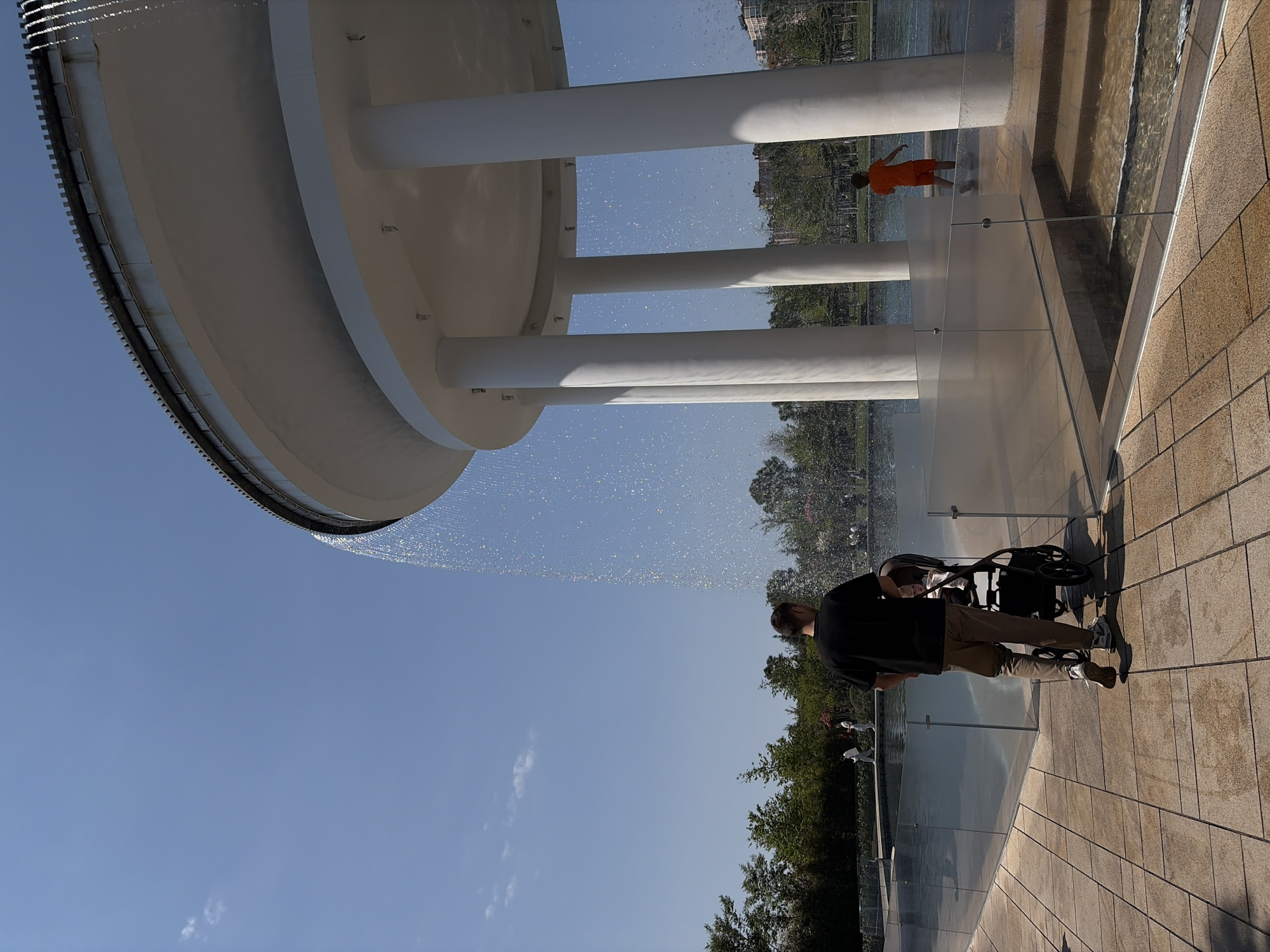 The image captures a scene at the Krasnodar Park in Krasnodar, Russia. A large, circular, architectural structure dominates the upper left, with water cascading from its edge. The sky above is clear blue, with a few scattered clouds.
A man wearing a dark shirt and light-colored pants is visible in the lower right, pushing a stroller. On the opposite side, further in the background, a person in orange clothing stands on the edge of a water feature.
The scene includes water elements, clear barriers, pavement, and vegetation, creating a blend of urban design and natural landscape.