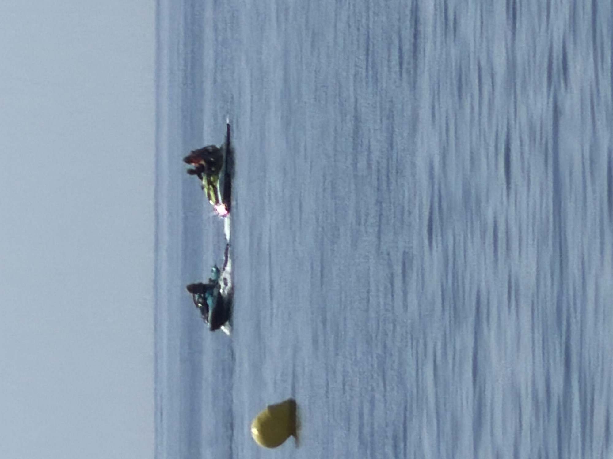 In Estepona, Spain, two jet skis are cruising on a calm, blue sea under a hazy sky. The first jet ski, on the left, is occupied by a single person wearing dark clothing. The second jet ski, to the right and slightly further back, carries two individuals. The person in front appears to be wearing a dark top and a helmet, while the person behind them is wearing a dark top and possibly an orange life jacket. There is a yellow buoy in the foreground to the left of the frame. The scene captures a recreational activity on the water.