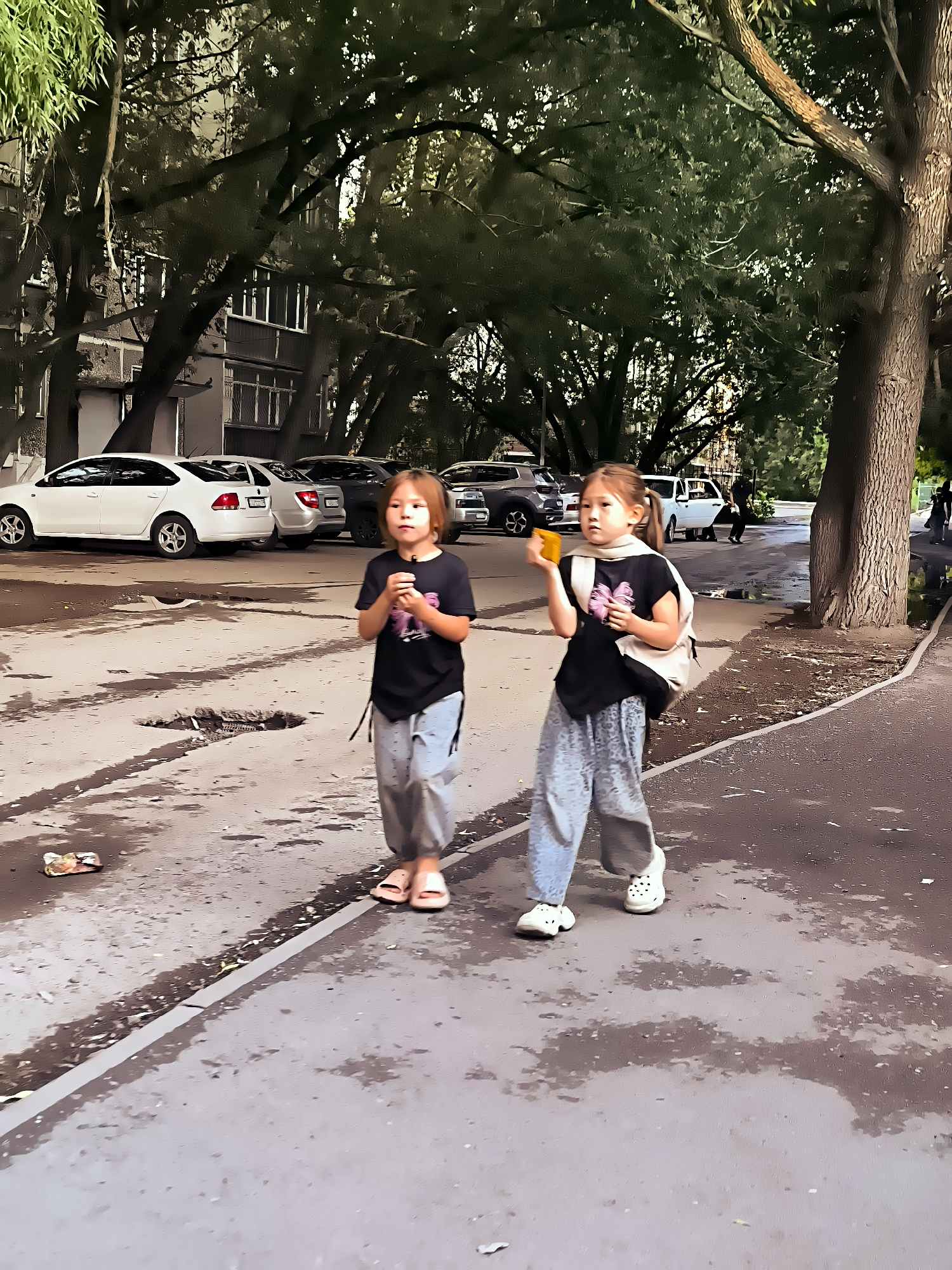 Two young girls, one with brown hair and the other with blonde pigtails, walk on a paved area outside. They are wearing t-shirts and loose-fitting pants. The girl on the left is holding a dark object, possibly a toy or food, near her mouth. The girl on the right has a light-colored backpack slung over one shoulder and is holding a yellow object in her hand. The ground is wet in patches, suggesting recent rain or puddles. In the background, several cars are parked in front of a multi-story building, and trees with dense green foliage line the area. The overall setting appears to be a residential courtyard in the city of Qarağandı, Kazakhstan.