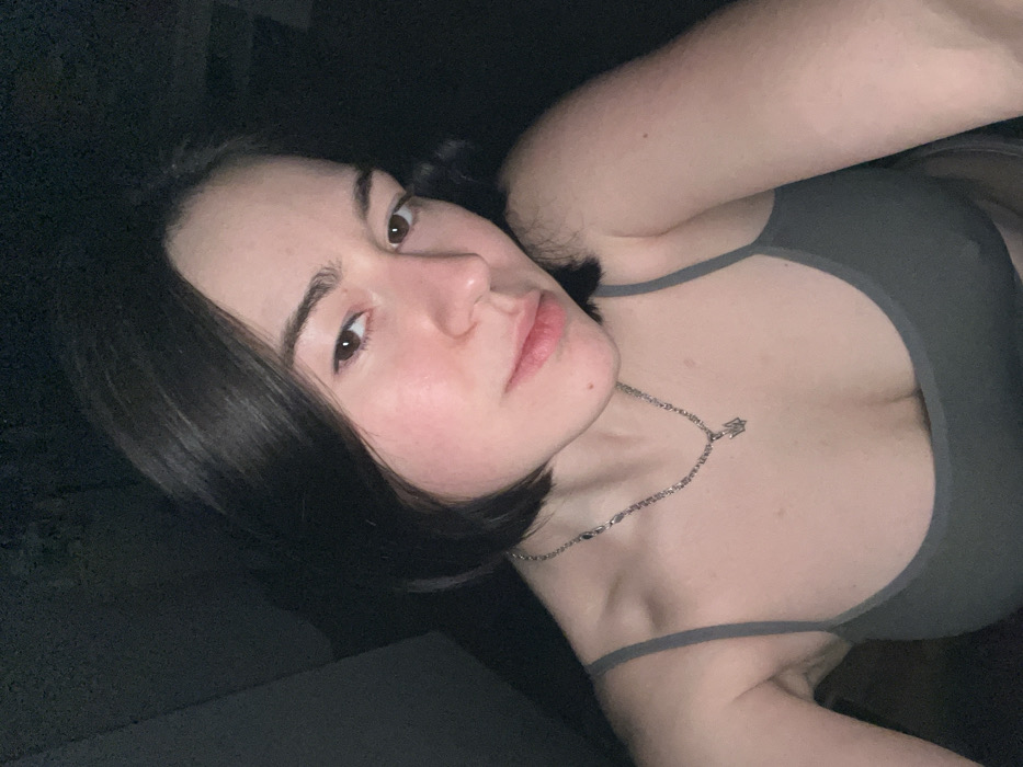 A woman with shoulder-length dark hair gazes at the viewer. She wears a gray tank top and a silver chain necklace with a pendant. Her skin is fair, and her expression is neutral. The lighting is dim, casting shadows around her face and body. The composition is at an angle, creating a dynamic and intimate perspective.