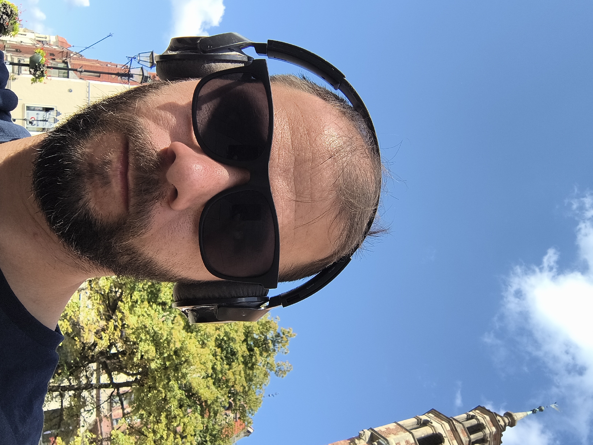 The image is a selfie of a man wearing black headphones and sunglasses. He has a beard and is looking directly at the camera. The background shows a clear blue sky with some clouds, trees with green foliage, and a building with a tower. The architecture of the tower suggests it might be a church or a historic building in a European city. The man is outdoors, likely in a public space. Based on the distinctive architecture of the building in the background, it appears to be St. John's Church and Cathedral in Toruń, Poland.