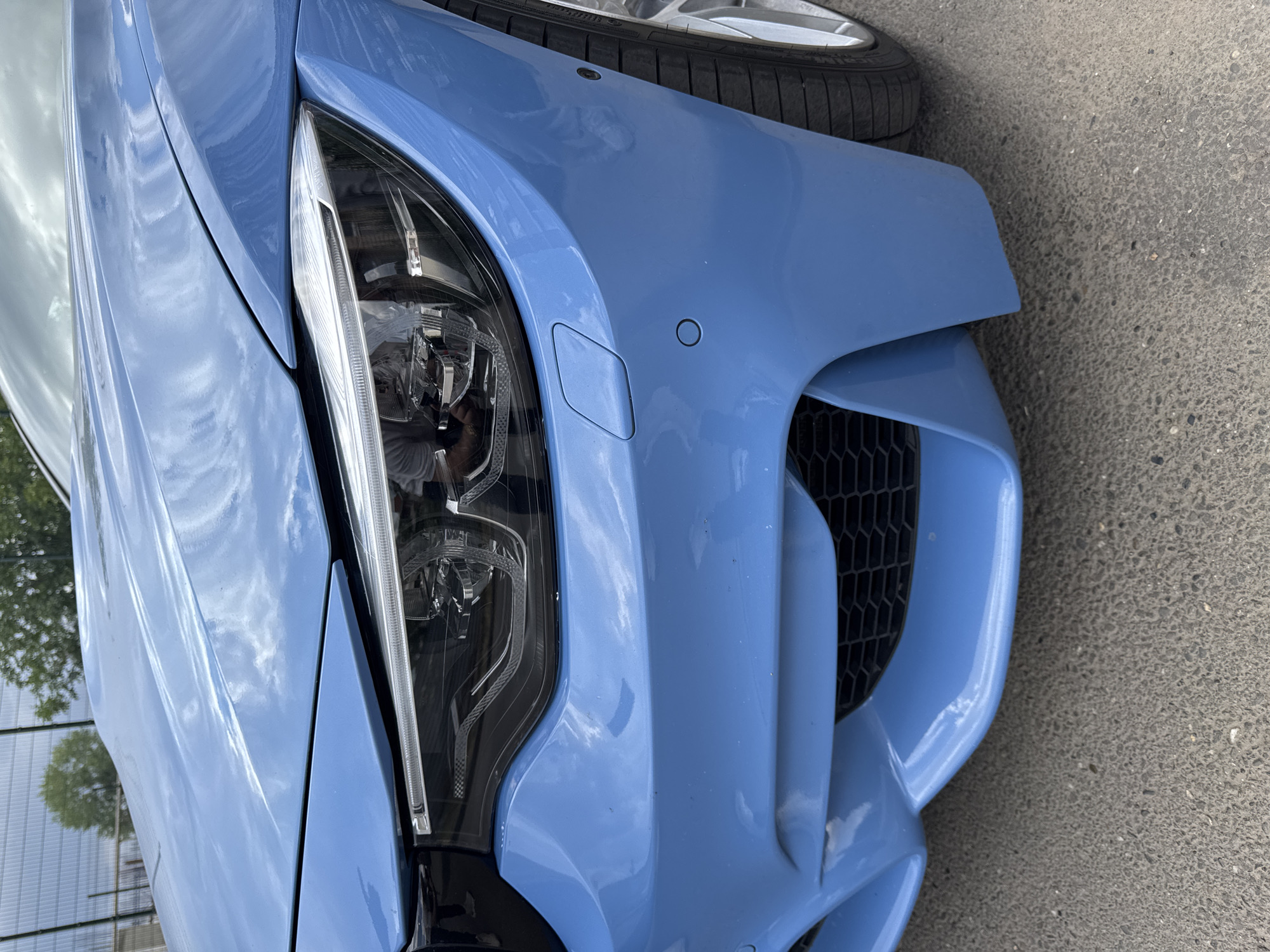 The image shows a close-up, eye-level, frontal view of a blue car. The car's headlight and bumper are clearly visible, with a black grille on the lower bumper. The car appears to be parked on an asphalt surface, possibly a street or parking lot. In the background, a chain-link fence and some trees can be seen, suggesting an outdoor setting. The sky is visible at the top of the image, with clouds reflecting on the car's polished surface. There is no indication of the car's location within the city of Krasnodar, Russia.