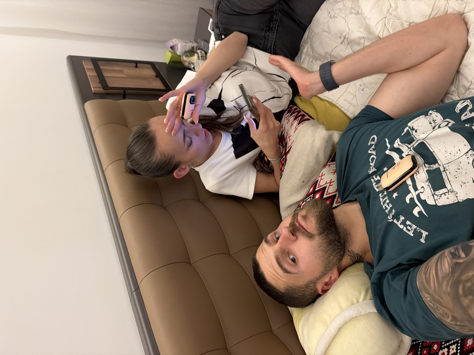 A man and a woman are lying on a bed, both looking at their phones. The man is in the foreground, wearing a teal t-shirt with a graphic print. He has a beard and tattoos on his arm. The woman is behind him, wearing a white t-shirt with a black graphic. She is holding her phone and a small pink object. The bed has a brown leather headboard and pillows with patterned covers. A nightstand with a lamp and a box is visible in the background. The room appears to be a bedroom in an apartment in Sofia, Bulgaria.