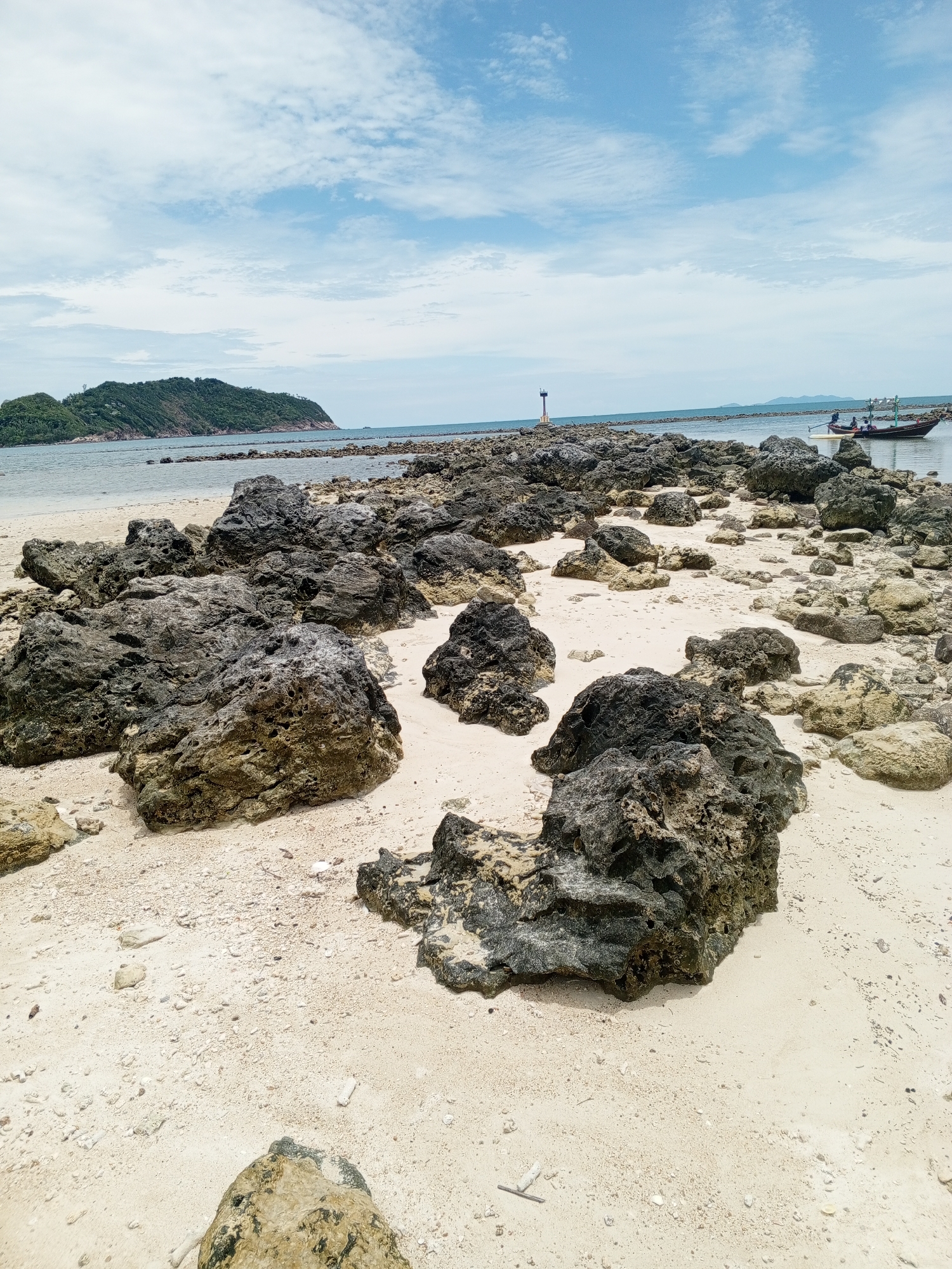 The image captures a coastal scene in an unknown location. The foreground is dominated by large, dark-colored rocks scattered across a white sand beach. In the middle ground, a stretch of rocks extends into the water, potentially forming a natural breakwater. In the background, a small, lush island sits on the horizon under a sky with scattered clouds. A small boat is anchored near the rocky breakwater, and a navigation marker is visible in the distance.