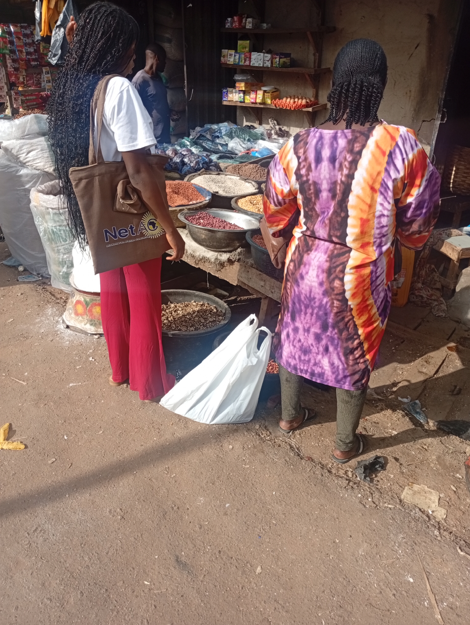 In Rafin Par, Nigeria, two women are seen at an outdoor market stall. The woman on the left, facing away from the camera, wears a white t-shirt and red pants, carrying a brown tote bag with a "NetAo" logo. The woman on the right, also facing away, wears a vibrantly patterned purple, orange, and yellow dress and dark green striped pants. They are standing near various sacks and bowls filled with grains and other food items, likely for sale. The ground is dusty and uneven, typical of an open-air market. Behind them, shelves are stocked with goods. A man can be seen in the background, partially obscured. The scene captures a moment of commerce and daily life within the market.