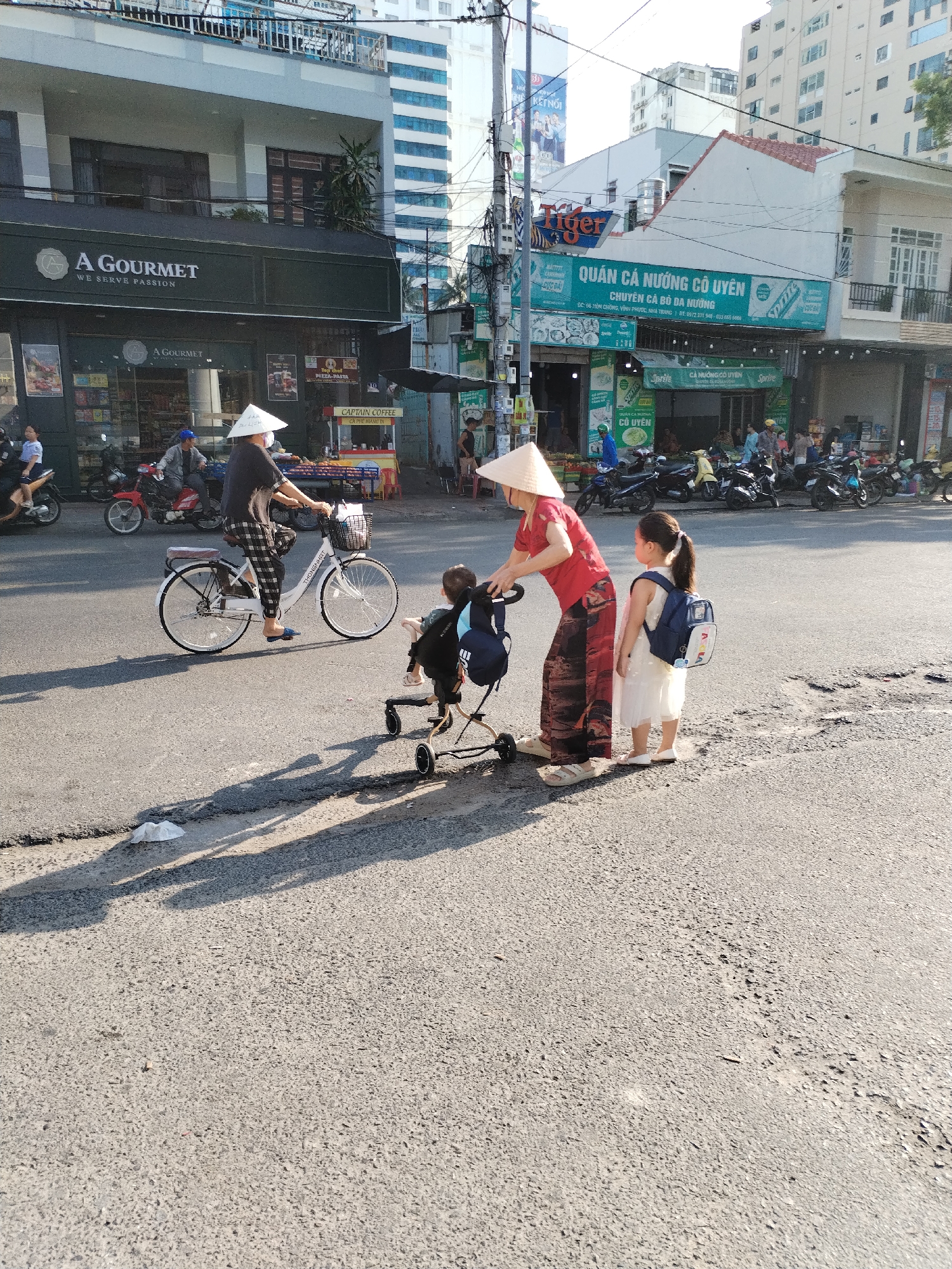 The image depicts a street scene in Bac Nha Trang Ward, Vietnam. An adult wearing a red shirt, patterned pants, a conical hat, and sandals is standing next to a stroller that contains a small child. Beside them stands a young girl in a white dress and a blue backpack. To their left, a person wearing a conical hat rides a white bicycle. In the background, there are several motorbikes parked along the street in front of shops and restaurants, including "A Gourmet" and "QUÁN CÁ NƯỚNG CÔ UYÊN." Traffic and buildings are visible further down the street. The lighting indicates it is daytime, with long shadows stretching across the asphalt.