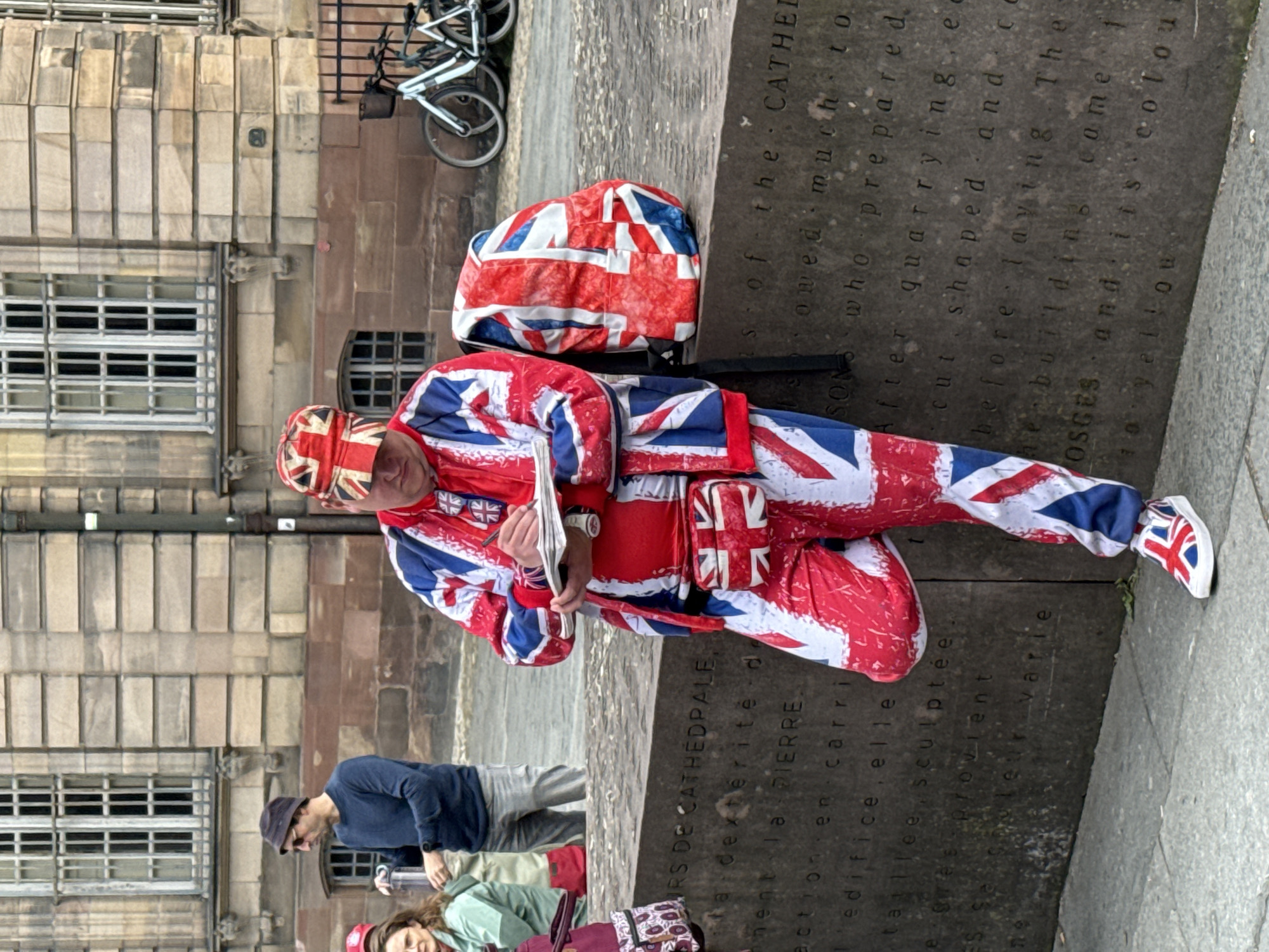 The image shows a man wearing a full-body suit, hat, and shoes all patterned with the British Union Jack flag, kneeling next to a stone block inscribed in French. The man is holding what appears to be a pamphlet or folded paper in his hands. A backpack with the same Union Jack pattern rests on the stone block near him.
In the background, a building with several windows is visible, along with a bicycle parked against a fence. To the left of the man, other people are passing by, including one wearing a blue shirt and another with reddish-pink hair and a green shirt.
The inscription on the stone block reads "COURS DE CATHEDPALE" at the top, indicating the location is likely near the Strasbourg Cathedral, in Strasbourg, France. The inscription continues with phrases related to stone and building materials, suggesting the block is a commemorative or informational marker related to the construction of the cathedral.