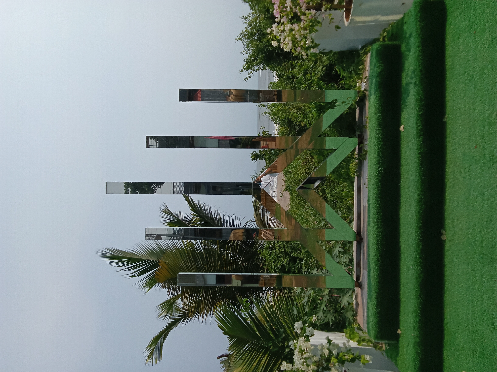 A vertical, outdoor shot presents a scene in Ashvem, India, with elements of nature and architecture. The clear sky in the background transitions into a view of the ocean. A palm tree is positioned in the foreground, along with plants and white flowers. A green, fence-like structure is present, possibly acting as a decorative piece or a barrier. Three reflective, rectangular panels are positioned above the fence and foliage, mirroring the surroundings and creating distorted reflections of buildings. Steps covered in green material are visible on the right, adding to the architectural details. The scene is set outdoors, capturing the environmental and structural components of a location in Ashvem.