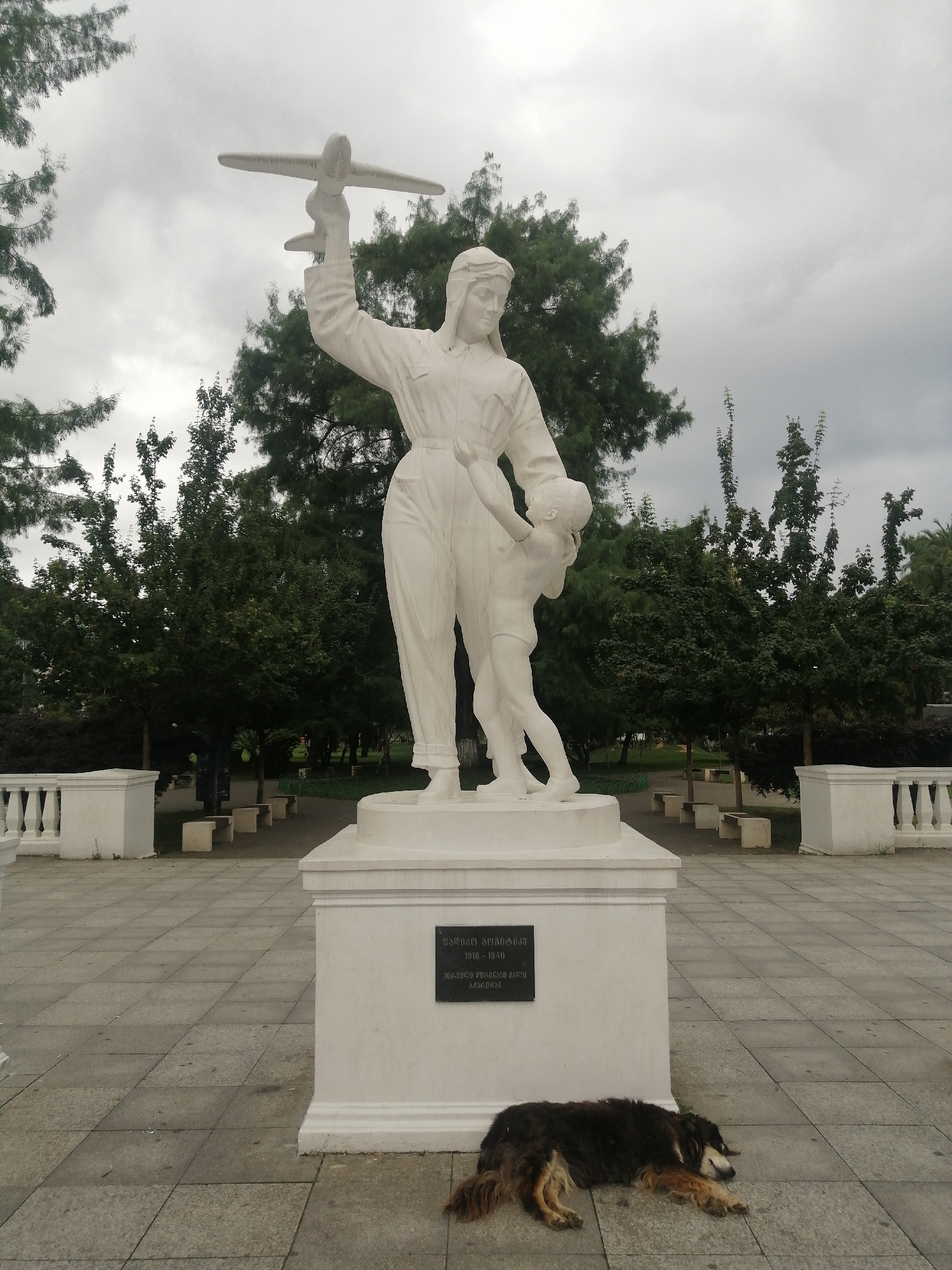 The image shows a white marble statue of a woman holding a model airplane, with a child clinging to her. The statue is on a pedestal with a plaque that appears to be in Georgian script. A dog is sleeping in front of the statue. The statue is located in Batumi, Georgia, in front of the Batumi Archaeological Museum. The sky is overcast, and there are trees and benches in the background. The ground is paved with large square tiles.