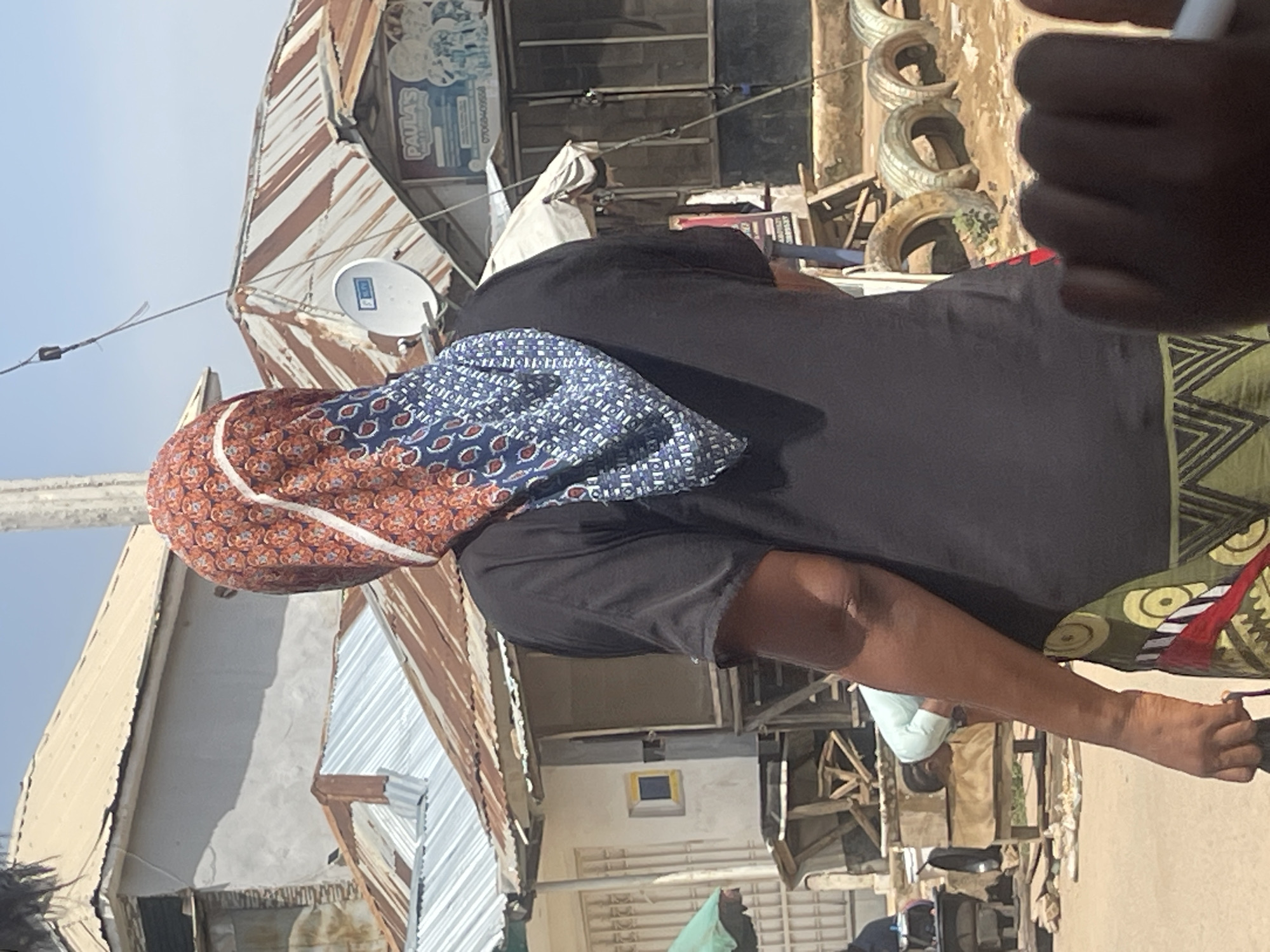 In Abuja, Nigeria, a person stands with their back to the camera, wearing a black t-shirt and a headscarf. The headscarf features a pattern of red and blue with white accents, and is draped over the person's head and shoulders. The person is also wearing a skirt or trousers with a green and yellow patterned design. In the background, there are several buildings with corrugated metal roofs, some of which appear weathered and rusty. A satellite dish is visible on one of the roofs. There is also a sign for "PAULA'S" which seems to be a business. In the foreground, there are several old tires stacked on the ground. The scene appears to be an outdoor market or a residential area with commercial activity.