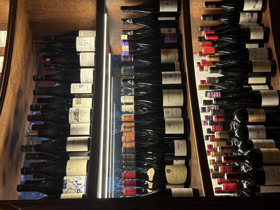 This image presents a detailed view of three stacked dark wood shelves densely populated with numerous bottles of wine within an indoor setting. Each shelf is illuminated by a recessed linear LED strip light positioned along its underside. The bottles are primarily dark-bodied, indicative of red wine, and feature diverse labels clearly displaying French appellations such as Saint-Joseph, Côte-Rôtie, Gigondas, Sancerre, Mercurey, and other Burgundy regional designations. The arrangement suggests a retail display or a wine storage area within a professional establishment, likely a caviste or restaurant cellar in Paris, France. No individuals or active interactions are present in the scene.