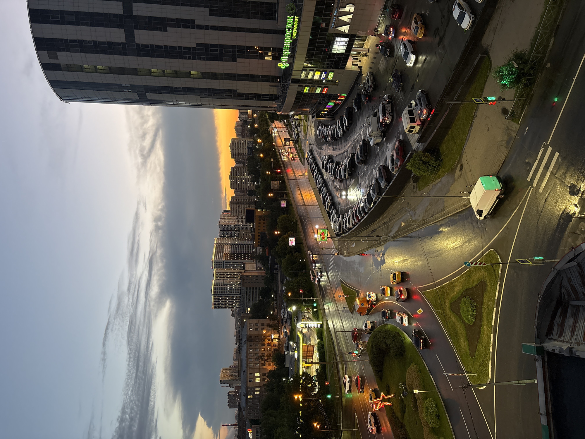 The image depicts an aerial view of a city intersection in Mytishchi, Russia, during twilight. The sky is a gradient of pale blue at the top, transitioning to hazy gray clouds and a bright orange sunset glow on the horizon. A large, modern, cylindrical building with a dark facade and vertical white and gray paneling dominates the right side of the frame. Several smaller, multistory residential buildings with illuminated windows are visible in the background.
The intersection is wet, indicating recent rain, with reflections of streetlights and vehicle lights shimmering on the asphalt. Numerous cars are navigating the roadway, including sedans, SUVs, and a few vans. One white van with a bright green illuminated roof is positioned near a pedestrian crossing. Traffic lights are visible, with some showing green signals.
A roundabout with green grass and a triangular landscaped area is situated in the foreground, with cars maneuvering around it. Adjacent to the roundabout, a gas station with yellow and white signage is lit up. A parking lot filled with cars is located at the base of the cylindrical building, with some vehicles parked in curved rows. Signs on the cylindrical building are visible, including "Перекресток" (Perekrestok) and "СУПЕРМАРКЕТ" (SUPERMARKET). Another sign on the lower portion reads "ТОРГОВО-ОФИСНЫЙ ЦЕНТР" (TRADE AND OFFICE CENTER) with "АЛЬТ" (ALT) underneath. Trees line parts of the roadway and surrounding areas. The overall scene suggests a bustling urban environment in the early evening.