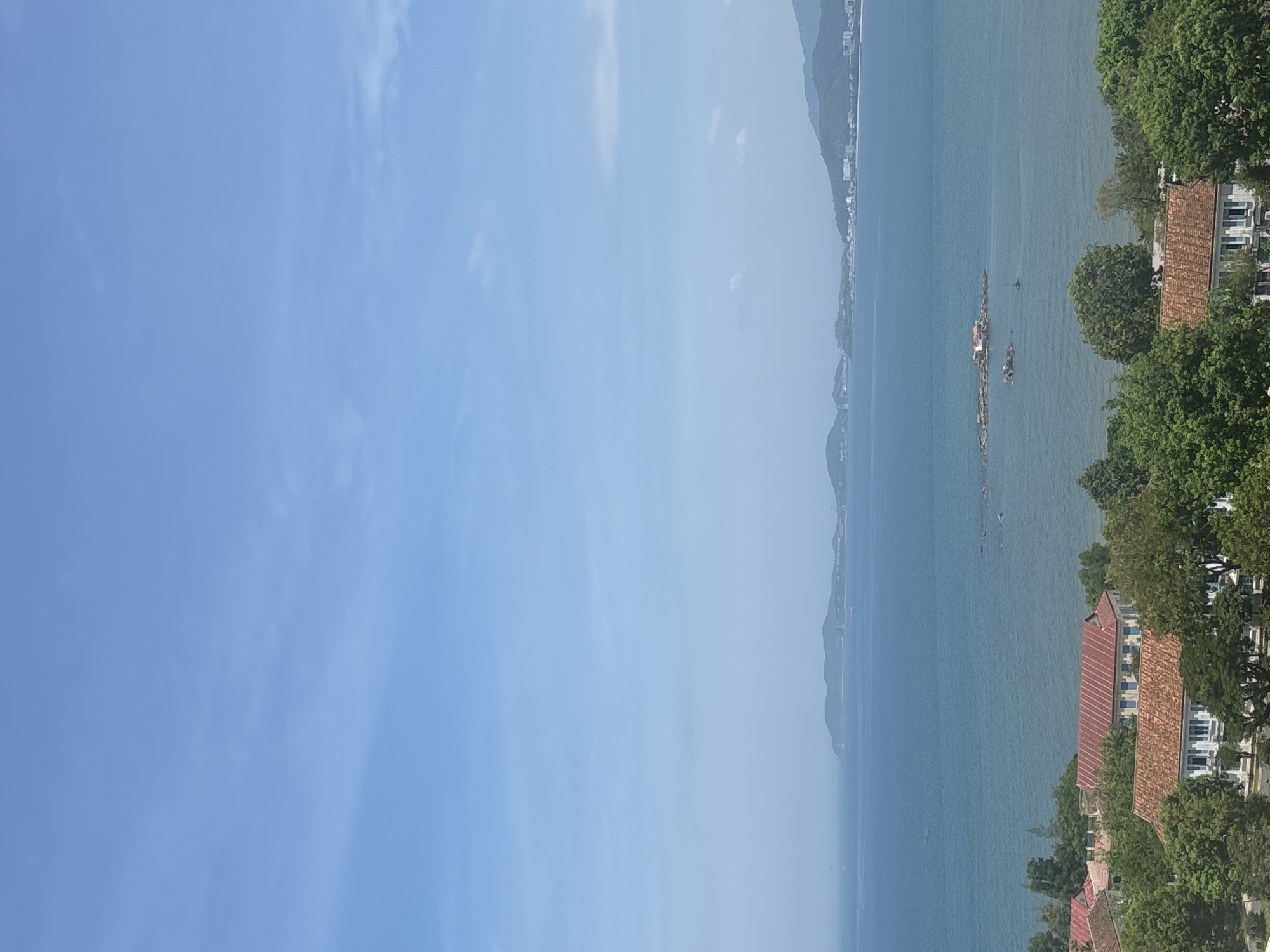 A high-angle, full shot captures a tranquil coastal scene under a bright, clear blue sky with scattered wispy clouds. The vast expanse of the ocean dominates the upper two-thirds of the frame, its surface a deep, calm blue. In the middle distance, a small rocky outcropping emerges from the water, featuring a single, small building with a red roof. A faint, distant coastline with rolling hills and scattered buildings can be seen on the horizon. In the foreground, a dense canopy of lush green trees partially obscures a cluster of buildings. These structures have terracotta-tiled roofs and white walls, with some displaying visible windows. The architecture suggests a blend of traditional and possibly older European styles. The scene evokes a sense of peacefulness and natural beauty.