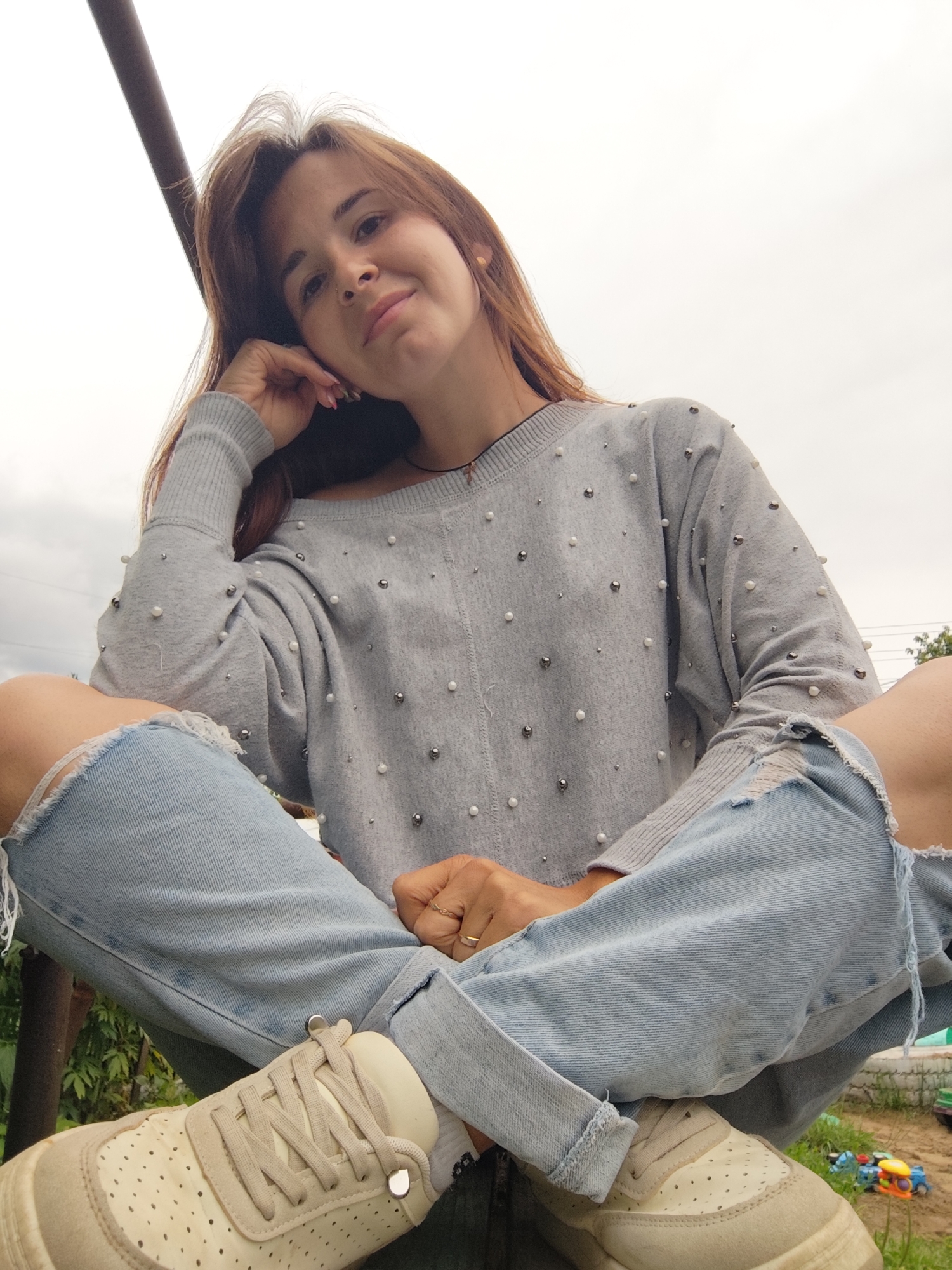 A woman with long, light brown hair is sitting outdoors, resting her head on her hand and looking at the camera. She is wearing a grey sweater adorned with pearls and metal studs, and ripped light blue jeans. Her legs are crossed, and she has on cream-colored sneakers. The background shows greenery and what appears to be a cloudy sky, suggesting a temperate climate. The setting is outdoors, possibly a backyard or a park. There are no immediate discernible landmarks linking the scene specifically to the city of Фоки, Russia.