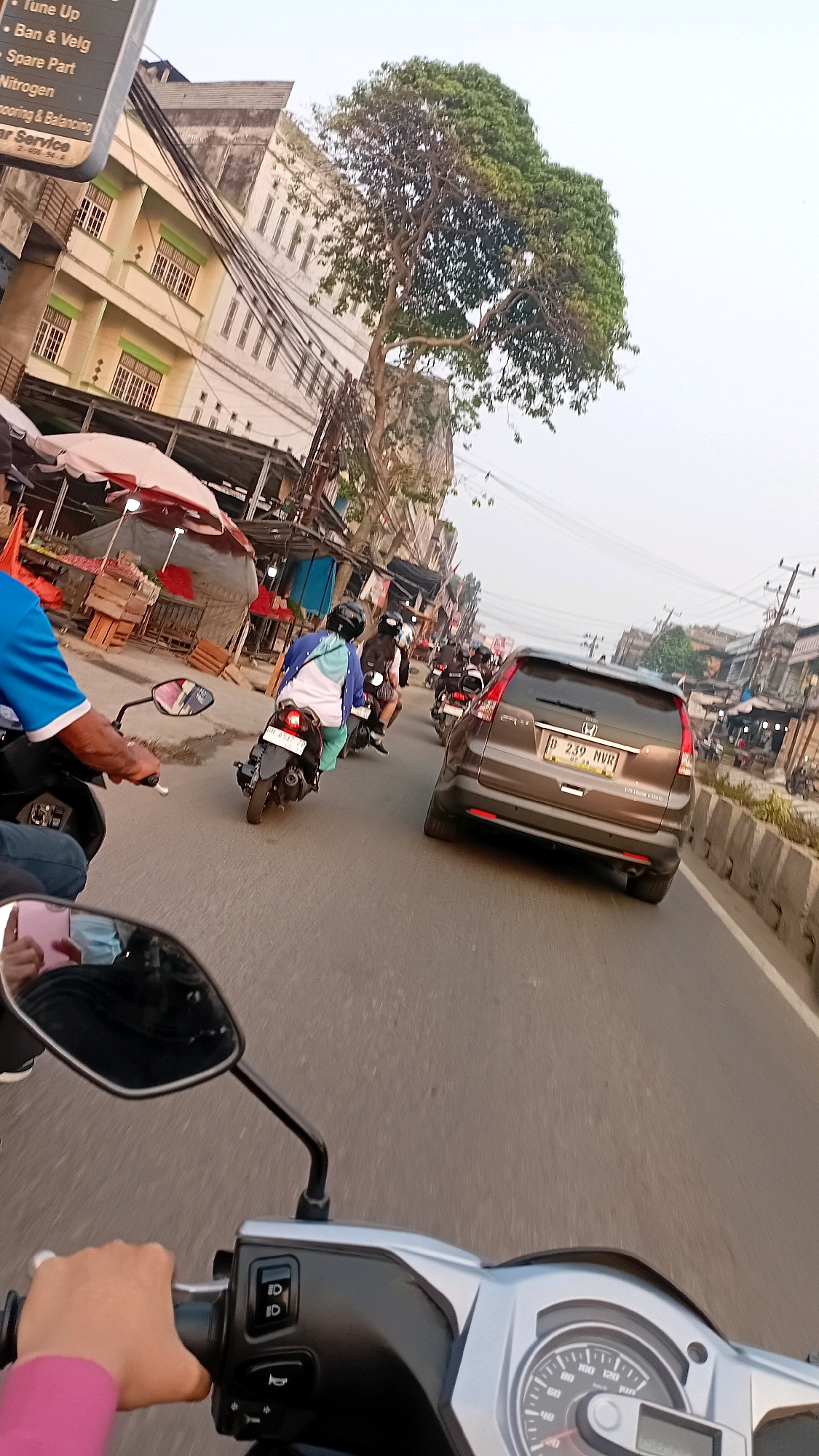 The image captures a street scene in Jambi, Indonesia, from the perspective of a person riding a motorcycle. The foreground shows the handlebars and part of the instrument cluster of the motorcycle, with a hand wearing a pink sleeve gripping the handlebar. In the mid-ground, a brown SUV with license plate B 239 MVR is visible, followed by several other motorcycles with riders wearing helmets. One motorcycle in the middle ground has a rider wearing a blue and white top. The background features buildings lining the street, some with multiple stories and balconies, and a large tree with dense green foliage. Street vendors with awnings are also present along the roadside, with wooden crates stacked behind them. Power lines are strung across the scene. The overall atmosphere suggests moderate traffic on a city street during the daytime.
