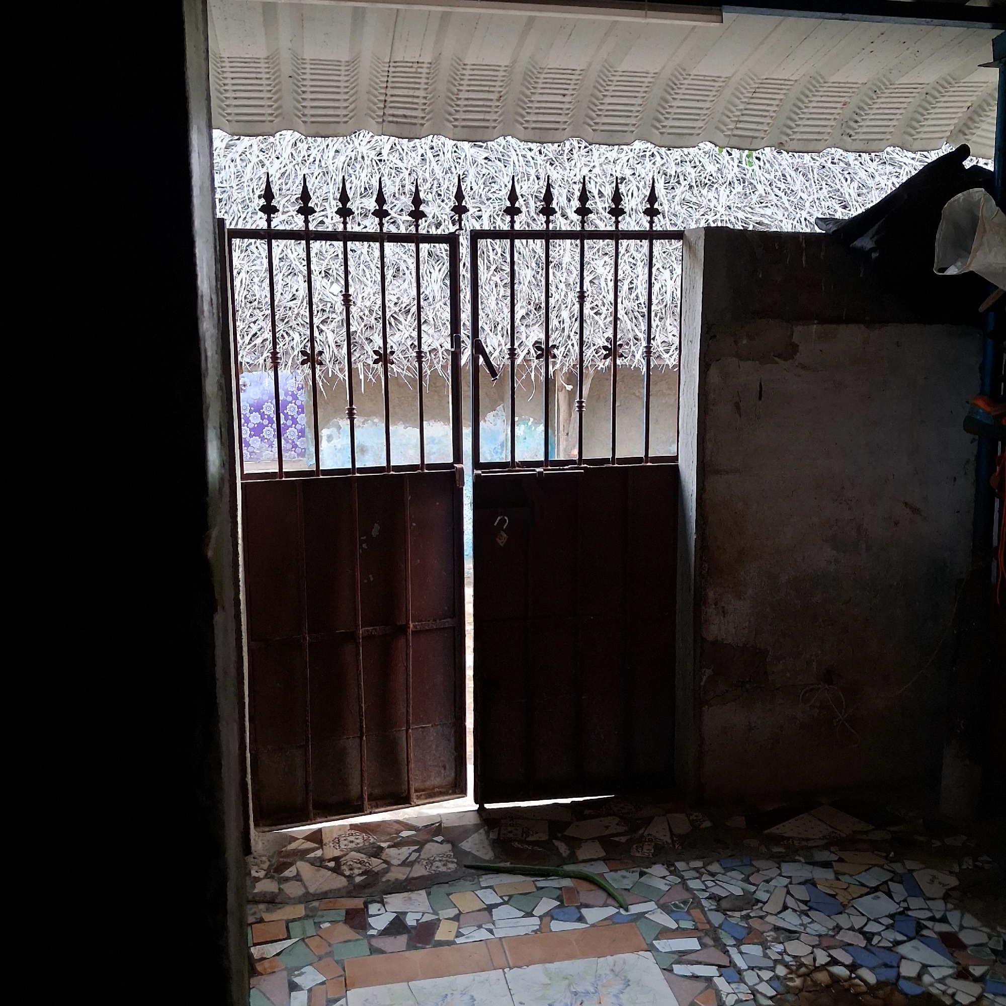 The image depicts a dark, enclosed outdoor space, likely a courtyard or entryway, in Nagapattinam, India. Two large, rusted metal gates stand open, revealing a bright, cluttered background filled with dry vegetation. The gates are topped with decorative iron spikes. The ground is covered in a mosaic of broken tiles in various colors and patterns, with some areas of intact tiles. To the right of the gates, a rough, textured wall is visible, appearing to be made of concrete or stone. Shadows are cast across the scene, indicating a light source from above and slightly behind the viewer. A long, green, flexible object, possibly a piece of plastic or vegetation, lies on the tiled ground in front of the gates. The overall impression is one of disrepair and neglect.