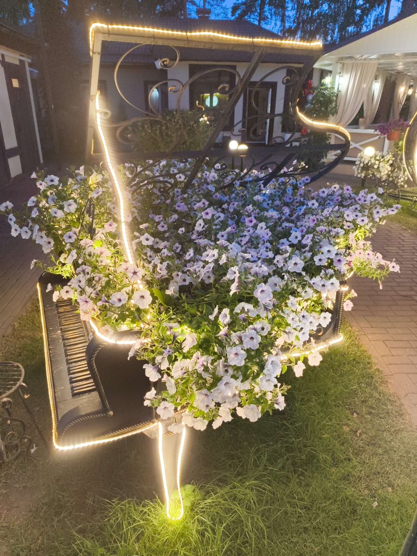 The image shows a decorative piano planter filled with numerous white and light purple petunias, illuminated by bright LED string lights. The piano planter is situated outdoors, with a brick pathway visible to the right and a grassy area in the foreground and to the left. In the background, a building with white curtains and a gazebo structure can be seen. The scene is lit by artificial lights, suggesting it is nighttime or dusk. There are no people visible in the image. The overall setting appears to be a garden or an outdoor seating area of a establishment in Alfimovo, Russia.