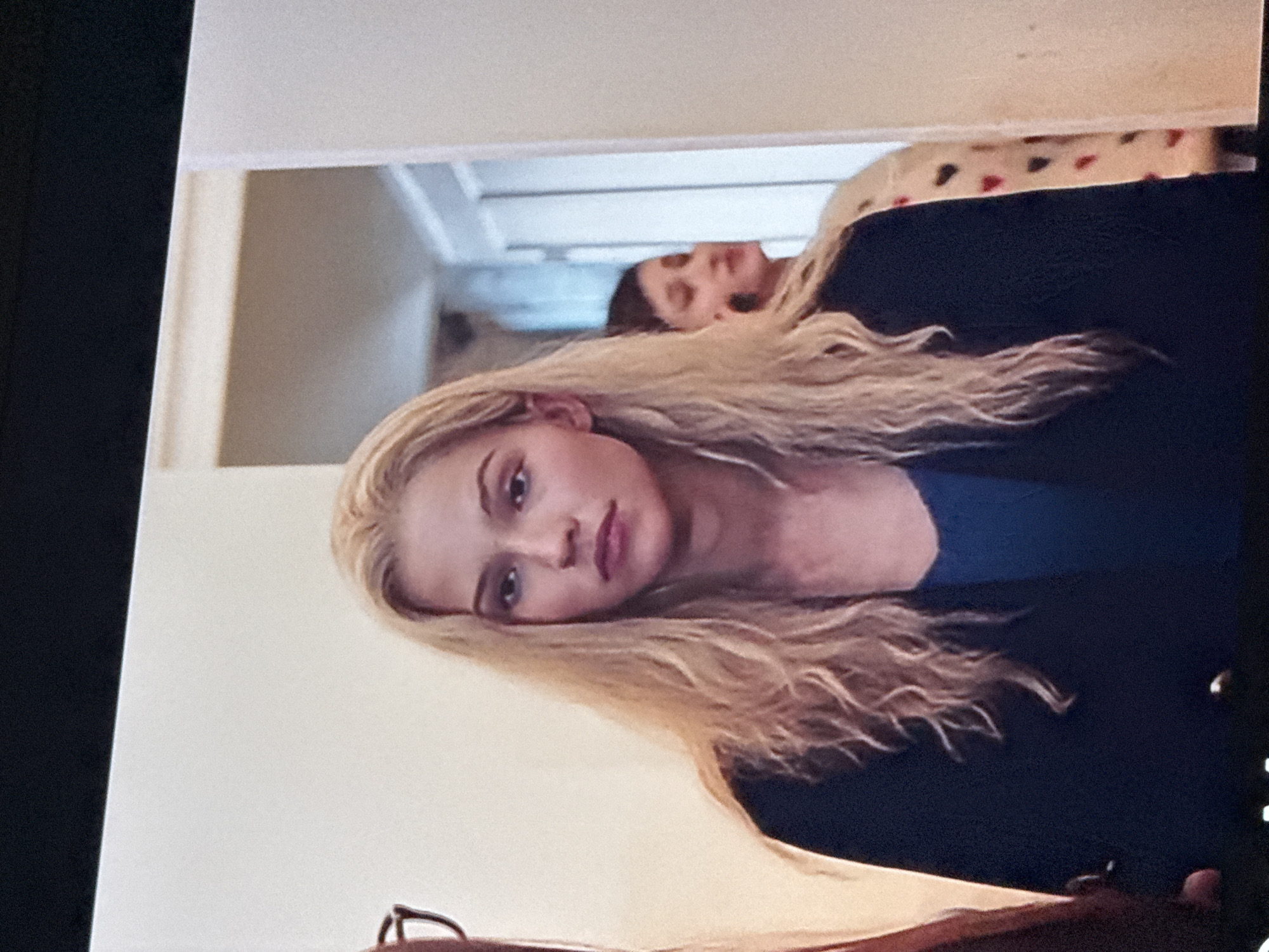 A woman with long, wavy blonde hair is in the foreground, wearing a dark blue top and a darker jacket. She is looking directly at the camera. In the background, through a doorway, a second person is partially visible lying on a bed covered with a blanket patterned with hearts. The environment appears to be indoors, potentially in a house or apartment. The background room is sparsely furnished. The location in the city of Mytishchi, Russia is not determinable from the image.