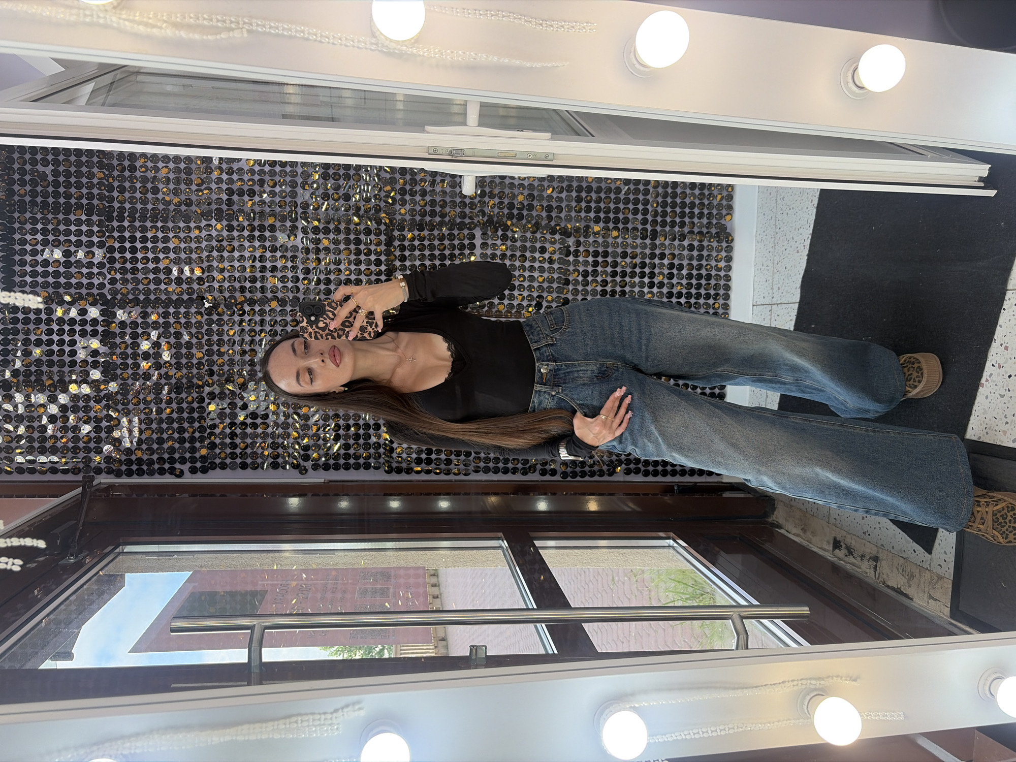 In a dimly lit indoor setting, a young woman stands facing a large mirror, taking a selfie with her smartphone. She is wearing a black long-sleeved top with lace trim, high-waisted blue wide-leg jeans, and leopard-print platform shoes. Her long, dark hair is down, and she is accessorized with rings and a necklace. The background behind her is a wall covered in black and gold sequins, creating a dazzling effect. To her left is a large glass door with a metallic handle, offering a glimpse of an outdoor environment with buildings and greenery. To her right, the mirror is framed by a vanity light fixture with several bulbs. The floor is tiled with a black and white pattern, and a black mat is placed in front of the mirror. The overall scene suggests a dressing room or a makeup studio, possibly in a retail or entertainment establishment.