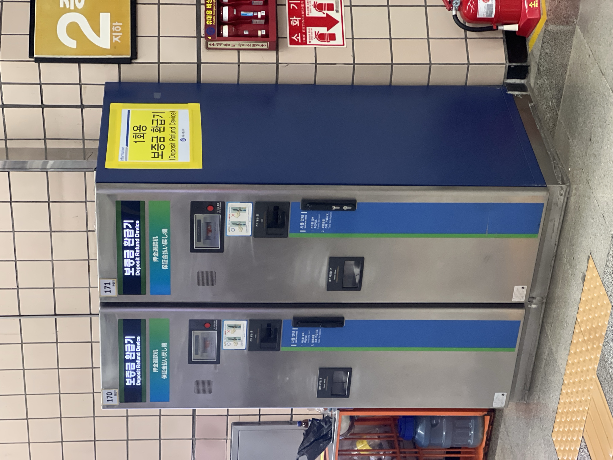 This media file shows a close-up view of two deposit refund machines installed in a subway station in Seoul, South Korea. The machines are silver with blue and green accents. Above the machines, a sign with the number "2" and the Korean word "지하" (meaning "underground") is visible, indicating the subway level. To the right of the machines, there is a red fire extinguisher mounted on a tiled wall, along with signage related to emergency lights and fire safety.
The machines are labeled in Korean and English as "Deposit Refund Device" and also include Japanese text. There are instructions on how to use the machines, which involve inserting a card and then retrieving the deposit. The scene appears to be indoors, likely in a public transportation setting, given the presence of the machines and the subway level indicator. The lighting suggests it could be daytime, but the exact time of day is not discernible. No people are visible in the image, and the overall atmosphere is functional and public. The machines themselves are the primary objects of interest, suggesting an activity related to returning items or deposits, possibly for reusable cups or containers, a common practice in South Korea. The detailed text on the machines and signs provides specific information about their function and usage.