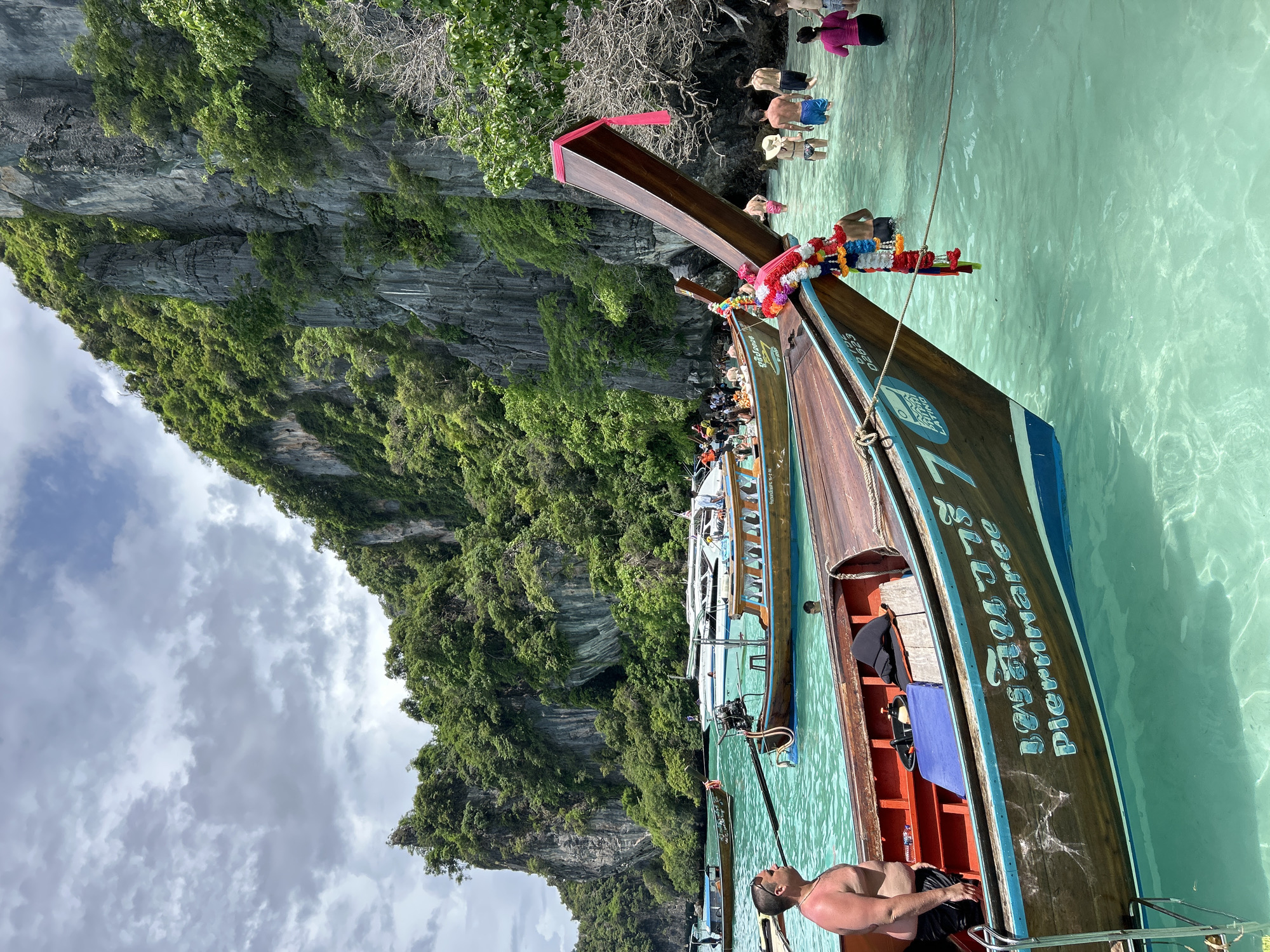 In Haad Hin Khom, Thailand, a body of clear, turquoise water is dotted with several wooden longtail boats. In the foreground, the bow of one such boat, adorned with a colorful garland of flowers and a pink ribbon, is prominently visible. A man in black shorts stands on the deck of this boat, looking to his left. Behind him, the interior of the boat reveals orange seats and a blue mat.
Further into the water, other longtail boats are anchored. One boat displays Thai script and the name "Piernnaree" in blue lettering. Several other boats, including a white motor yacht, are further out. A group of people, some wading in the water and others on boats, are visible in the midground. The background is dominated by a steep, lush green cliff face, characteristic of the region. The sky above is partly cloudy with a mix of grey and white clouds.