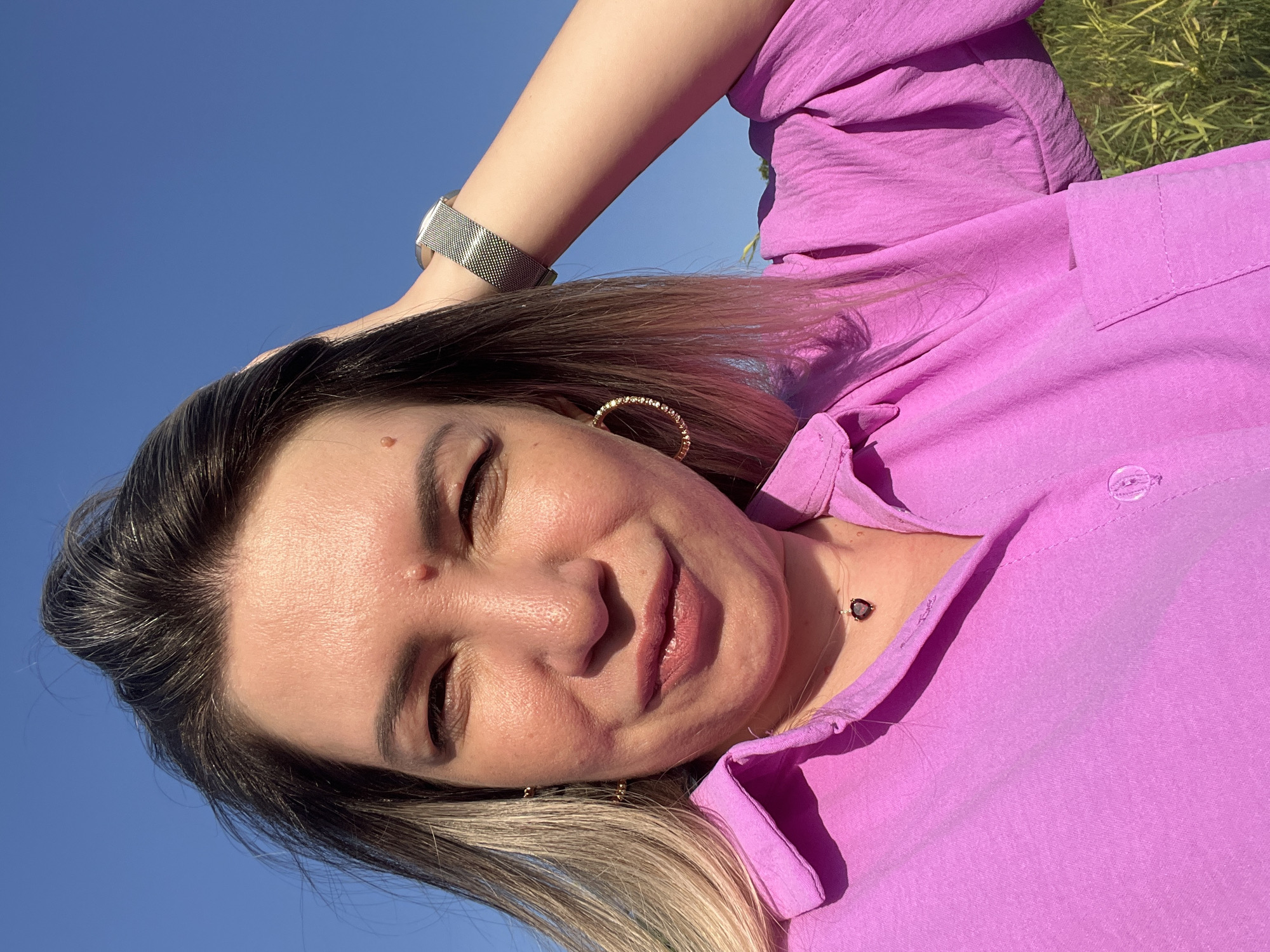 A woman is taking a selfie outdoors against a bright blue sky. She is wearing a pink button-up shirt and a necklace with a heart-shaped pendant. Her left hand is in her hair, and her right arm is raised behind her head, revealing a silver watch on her wrist. She has blonde and dark brown hair and is wearing hoop earrings. The background shows some green grass and foliage, suggesting a natural or park-like setting. There are no identifiable landmarks in the image that would indicate the specific location within Яново-Грушевский, Russia.