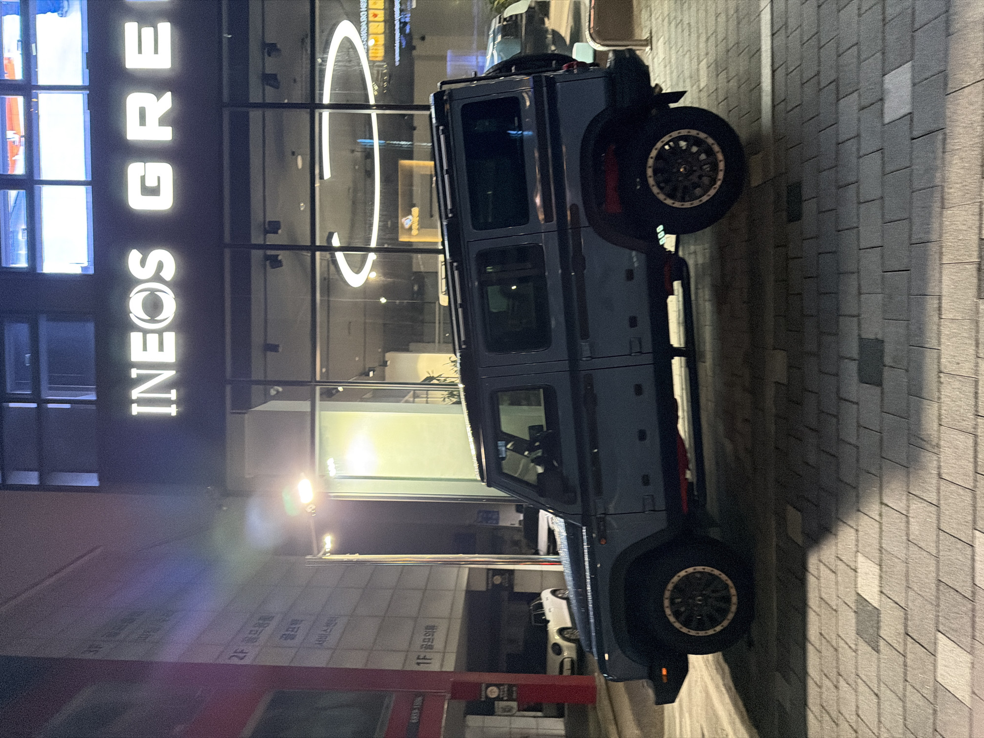 A dark blue, four-door SUV with a roof rack is parked on a brick-paved sidewalk at night. The SUV has custom wheels with a beige trim and red accents on the undercarriage. Behind the SUV, a white Mini Cooper is parked in a covered lot. In the background, a building with a large, illuminated sign reads "INEOS GREEN". The building has large glass windows with circular light fixtures visible inside. The environment appears to be an urban setting in Seoul, South Korea, likely a commercial district. The lighting is artificial and casts shadows on the pavement. There are no people visible in the image.