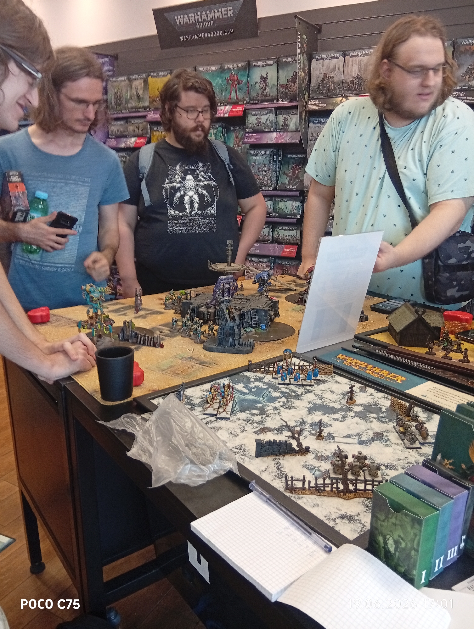 The image depicts a group of three men engaged in a tabletop wargame, likely Warhammer, in a retail store. The setting is identifiable as a Warhammer store due to the prominent branding and product displays visible in the background. Shelves are stocked with various Warhammer boxes, including "Warhammer 40,000," "Combat Patrol," and "Tyranids."
The individuals are focused on the game laid out on a table. One man, wearing glasses and a blue t-shirt, holds a smartphone in his hand. Another man, also with glasses and a beard, wears a black t-shirt with a graphic design and has a backpack slung over his shoulder. The third man, with long hair and glasses, is partially visible on the left. They are surrounded by game miniatures, terrain pieces, and dice, all indicative of a wargaming session.
On the table, a detailed game board with miniatures representing armies is set up. There's also a white plastic stand holding a sheet of paper, and to the right, some smaller boxed game elements. The overall atmosphere appears to be one of concentration and shared interest in the hobby. The lighting suggests an indoor environment, likely during daylight hours. A timestamp in the lower right corner indicates "19.06.2026 17:01," and "POCO C75" in the bottom left corner suggests the device used to capture the image.