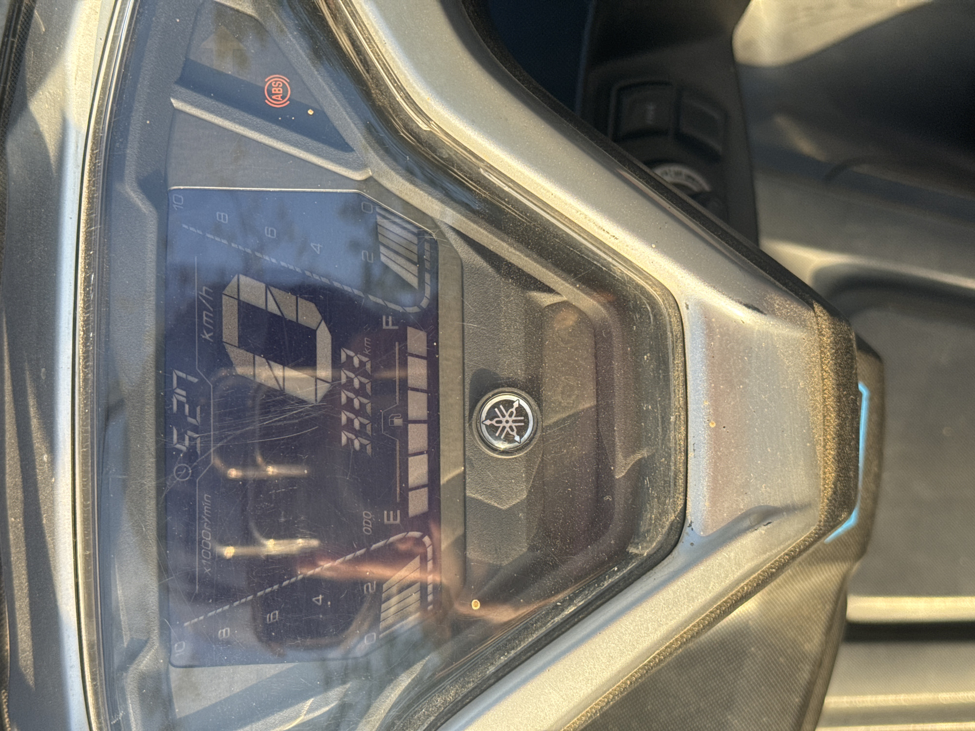 The image shows a close-up of a motorcycle dashboard. The odometer reads 33333 km. The fuel gauge is low, near the "E" for empty. The speedometer is showing a speed of 2 km/h. The RPM gauge reads approximately 5.27 x 1000 r/min. The ABS light is also visible in the corner. The background is blurred, suggesting the location is possibly Baan Chaweng, Thailand. The dashboard is dusty and has some scratches on the cover. The Yamaha logo is present on the dash.
