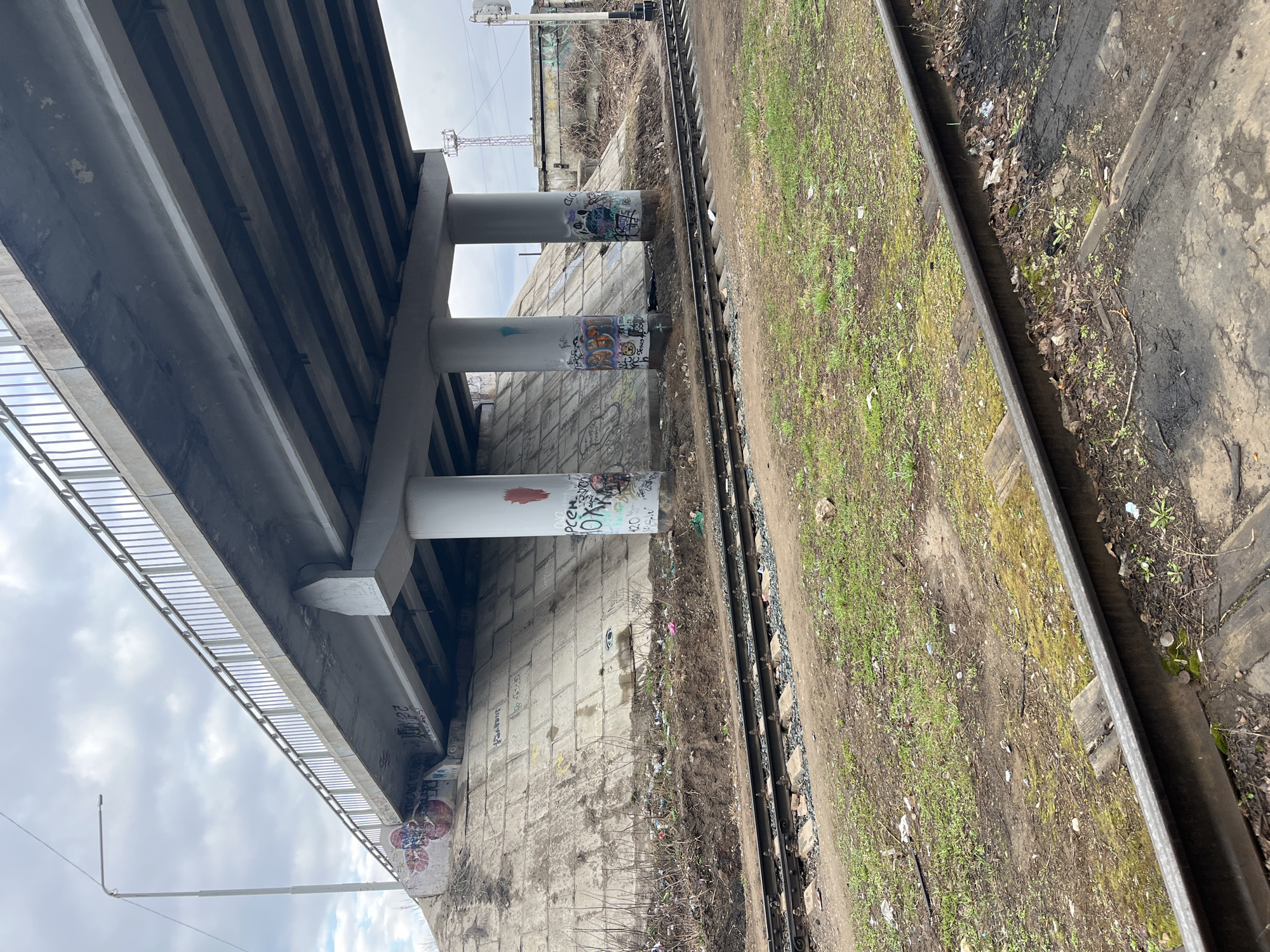 The image captures an elevated view beneath a large concrete overpass, showing its support structure and adjacent railway tracks under an overcast sky.
The main subject is a substantial concrete bridge or overpass, running diagonally across the upper portion of the frame. Its underside reveals numerous parallel concrete beams. Along the left edge of the bridge, a metal safety railing is visible. Three prominent, cylindrical concrete support columns (piers) extend from the bridge deck towards the ground. These columns are heavily covered in colorful graffiti tags and stylized lettering, with words like "PCE" and "OXN" partially visible on the upper column.
To the right of the columns, a set of railway tracks with wooden sleepers and gravel ballast stretches horizontally through the scene. The ground immediately surrounding the tracks and at the base of the bridge columns consists of dirt, sparse green grass, dry brown weeds, and scattered litter, indicating a somewhat neglected area. A retaining wall, constructed from large concrete blocks, is situated between the bridge columns and the railway line. This wall also bears faint, mostly illegible graffiti markings and numbers.
In the background, an overcast sky is visible, suggesting a cloudy day without direct sunlight. A slender metal utility pole or lamppost is seen extending upwards from the lower left, while a communication tower structure can be discerned in the distance on the right. No people or active events are present in the scene, which appears to be a static, utilitarian urban or industrial landscape. The overall impression is one of infrastructure and transit, with signs of human presence through graffiti and discarded items.