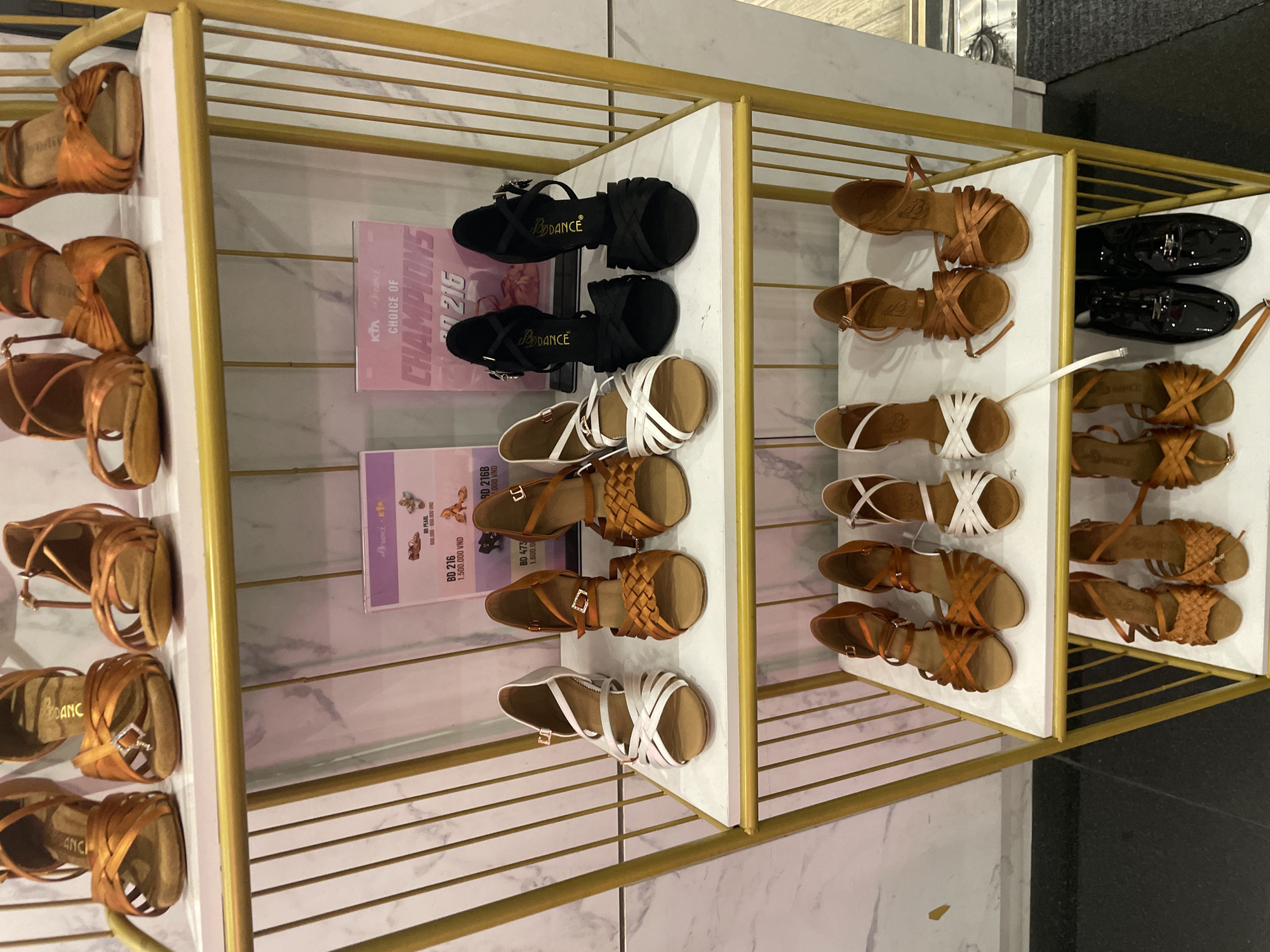 The image shows a retail display of ballroom dancing shoes in Thủ Đức, Vietnam. The shoes are arranged on gold-colored shelves against a marble-patterned wall. The display features various styles of dance shoes, including women's heeled sandals in tan and black, and some white strappy sandals. There are also two pairs of shiny black men's dress shoes on the top right shelf.
Several informational cards are visible among the shoes. One card, primarily pink, reads "KTADANCE CHOICE OF CHAMPIONS 2216". Another card, mostly purple, displays product codes and prices such as "BD PEARL 800.000-950.000 VND", "BD 216 1.500.000 VND", and "BD 473 1.800.0". The branding "BDANCE" is visible on several of the tan dancing shoes.
The overall scene is well-lit, suggesting an indoor retail environment. The focus is on the product display, with no people visible. The arrangement is neat and organized, indicating a professional sales setting.